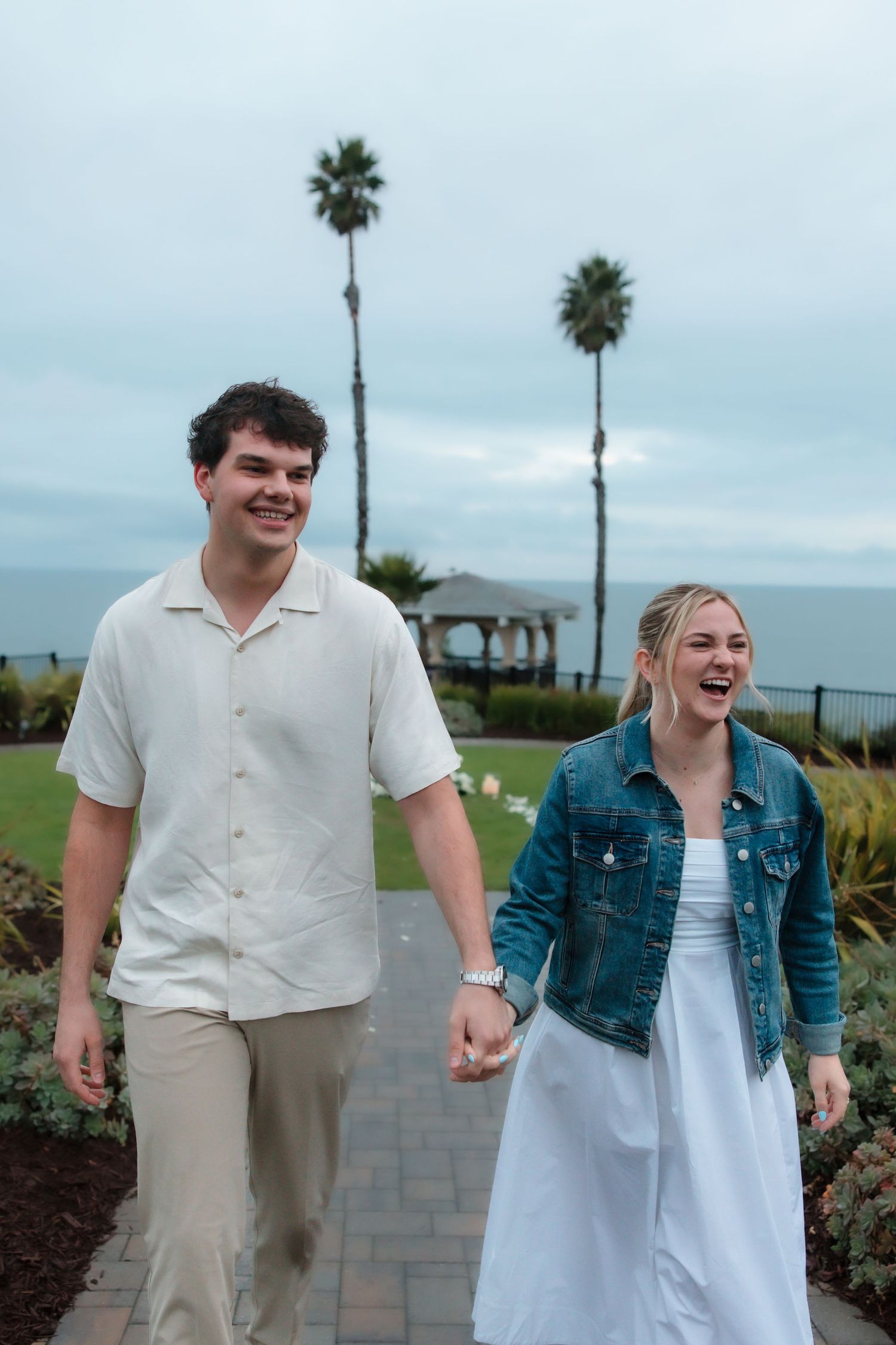 A smiling couple holds hands while walking along a garden path with palm trees and ocean views in the background.