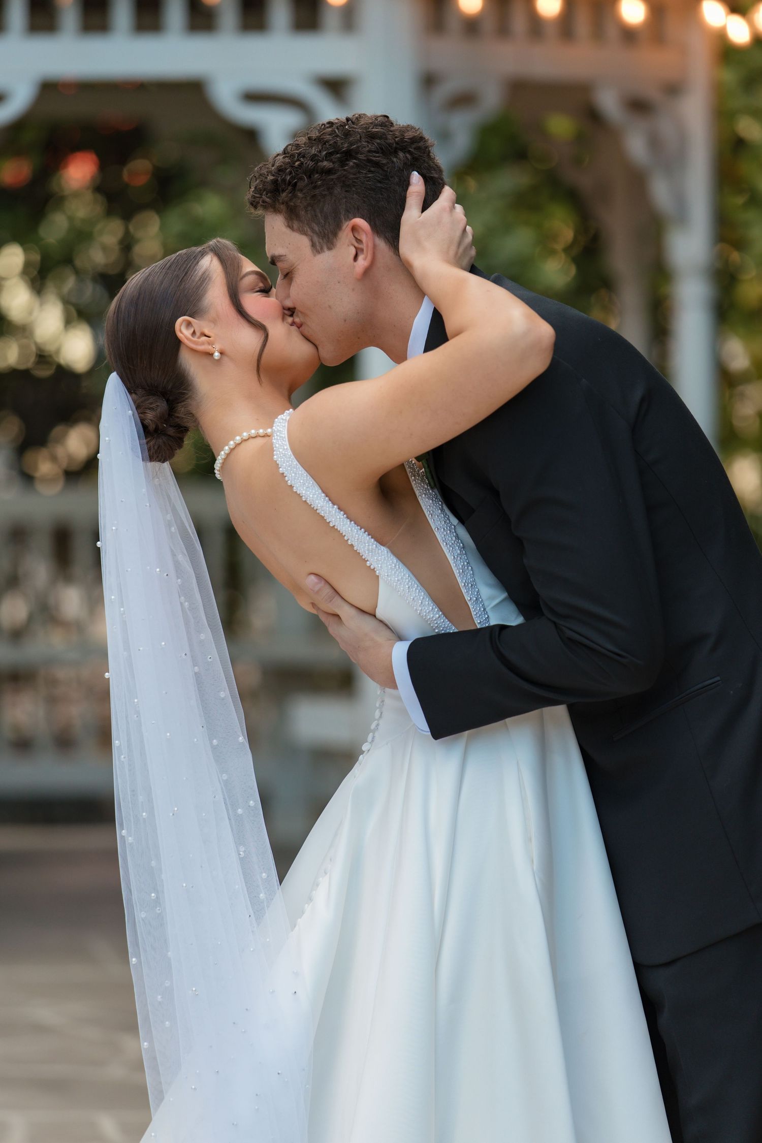 Bride and groom share a romantic first kiss closeup at outdoor gazebo wedding ceremony at dusk.