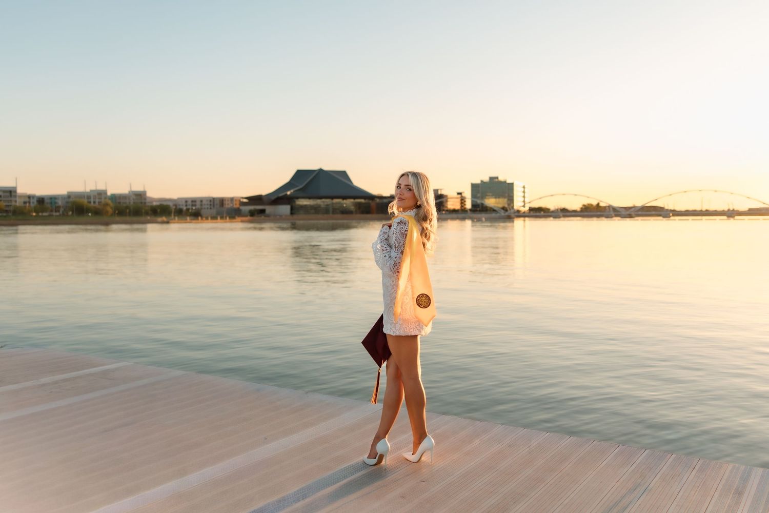 Blonde woman in white dress walking on a waterfront pier at golden sunset with city views.