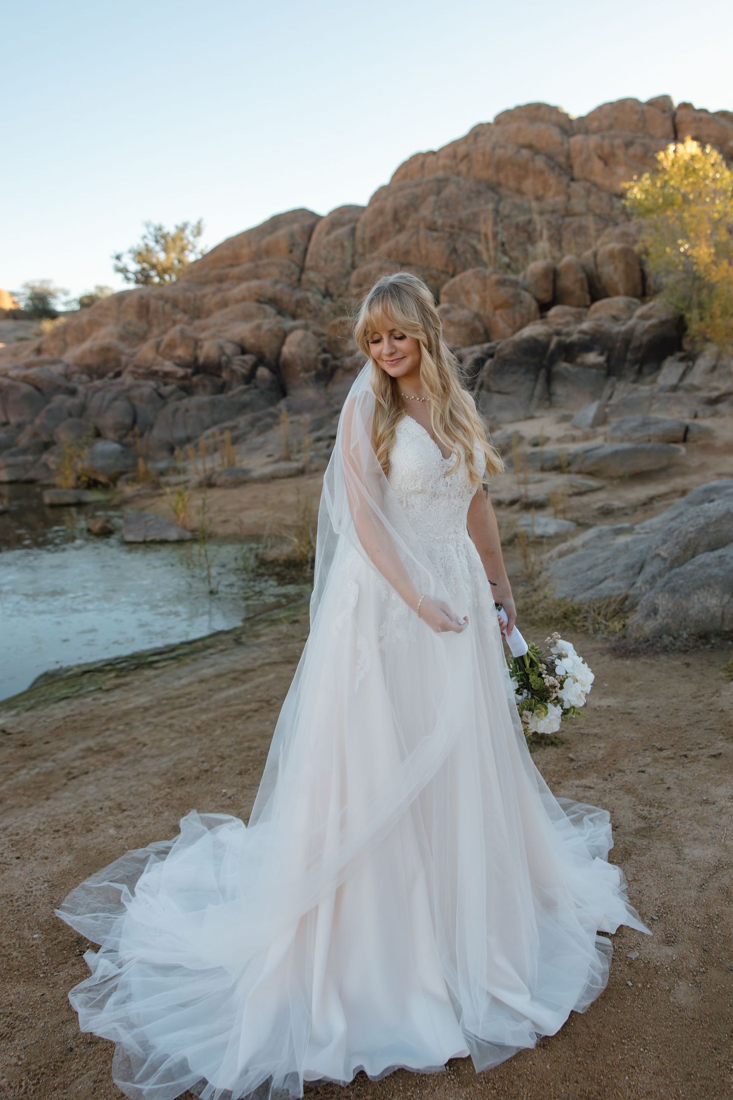 Radiant bride in flowing white gown with veil holds bouquet before scenic rocky lakeside backdrop.