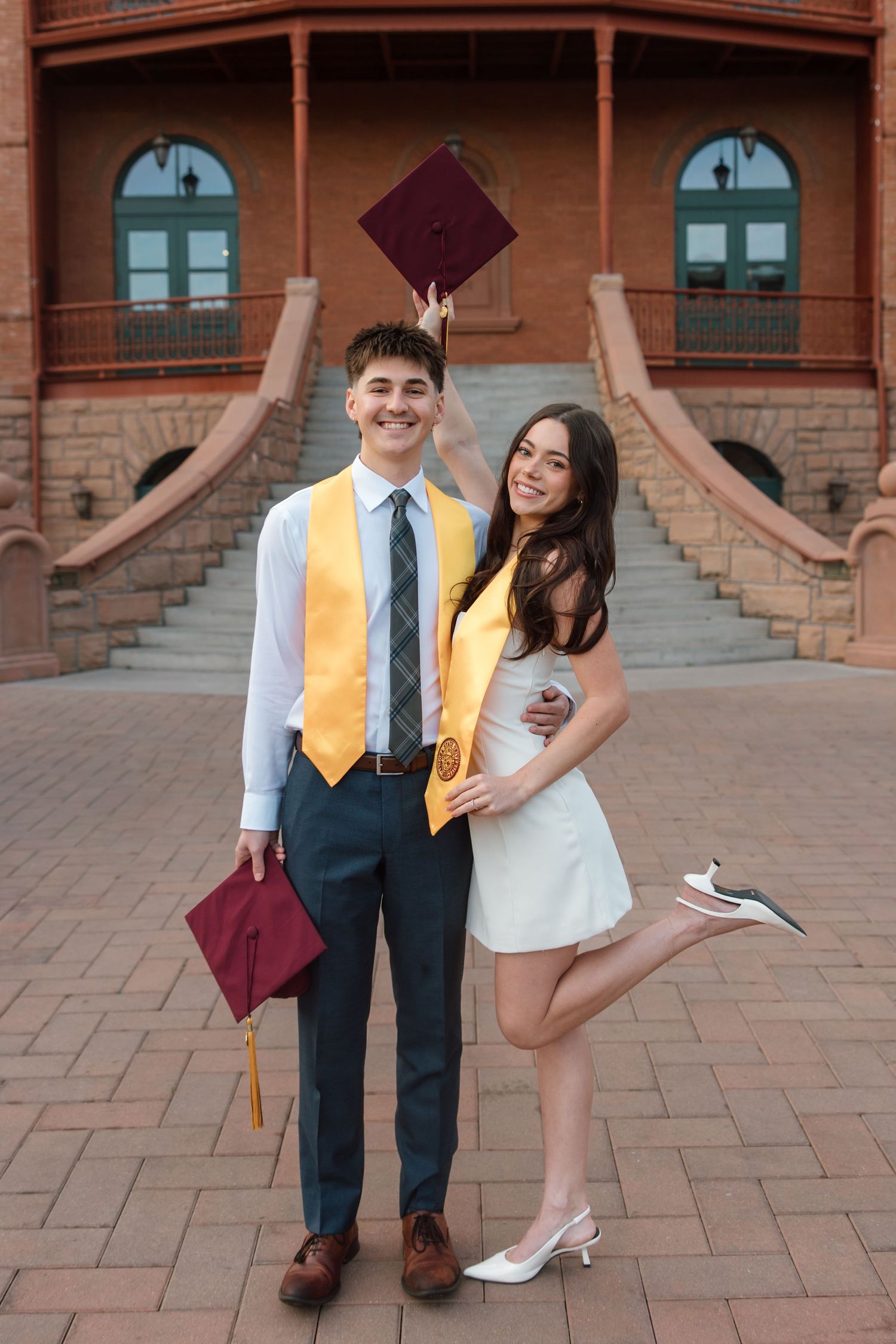 Smiling couple posing with graduation caps, wearing yellow stoles in front of a brick building.