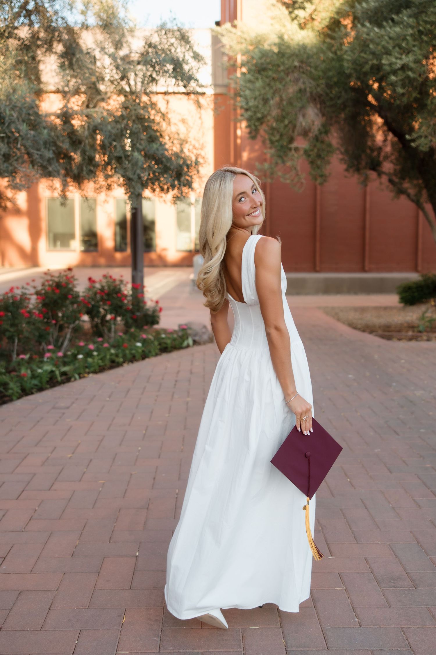 Blonde woman in white backless maxi dress holding maroon graduation cap outdoors.