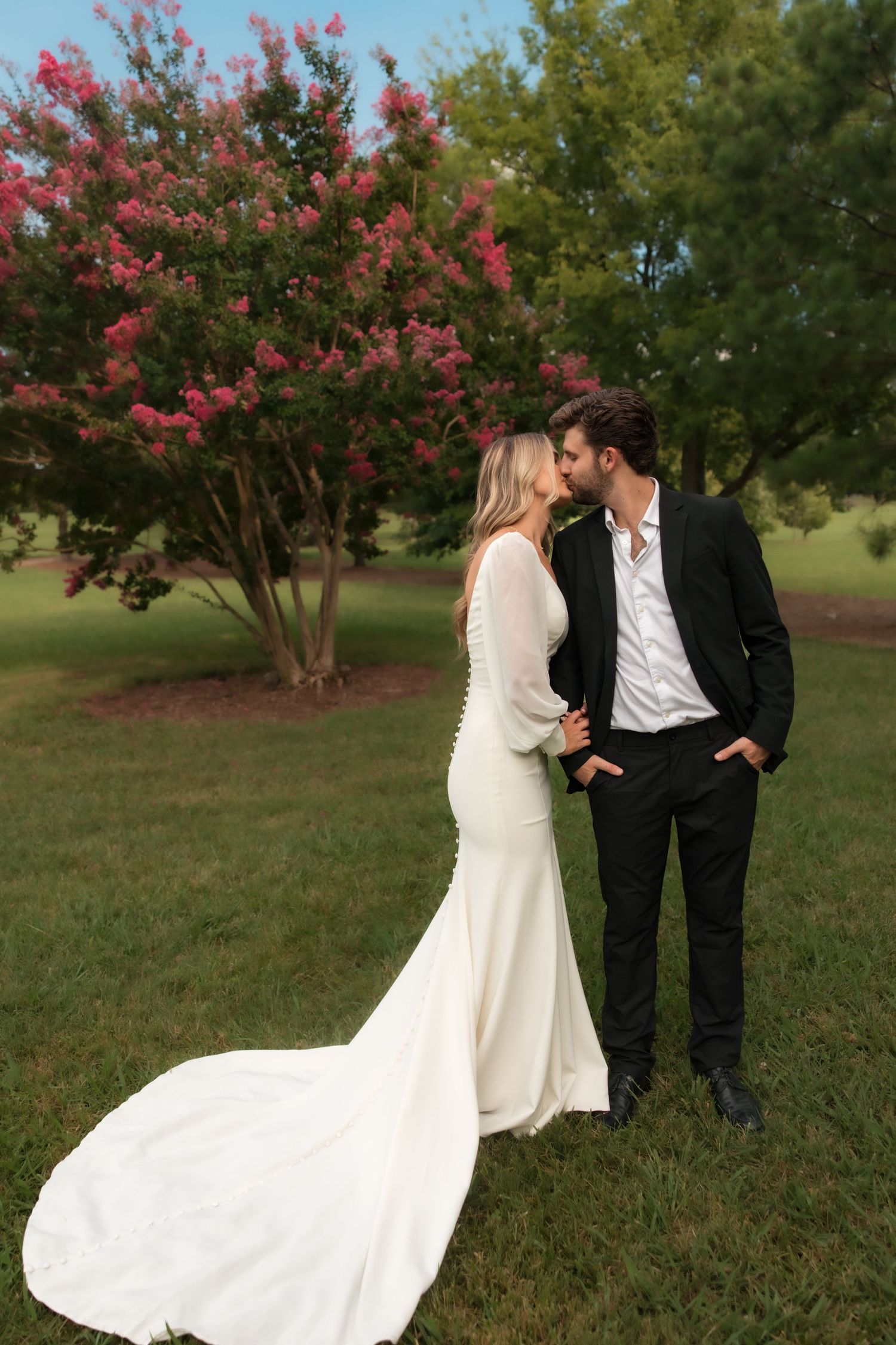 Bride and groom kissing outdoors in front of a blooming pink crape myrtle tree, bride in elegant white gown with train.