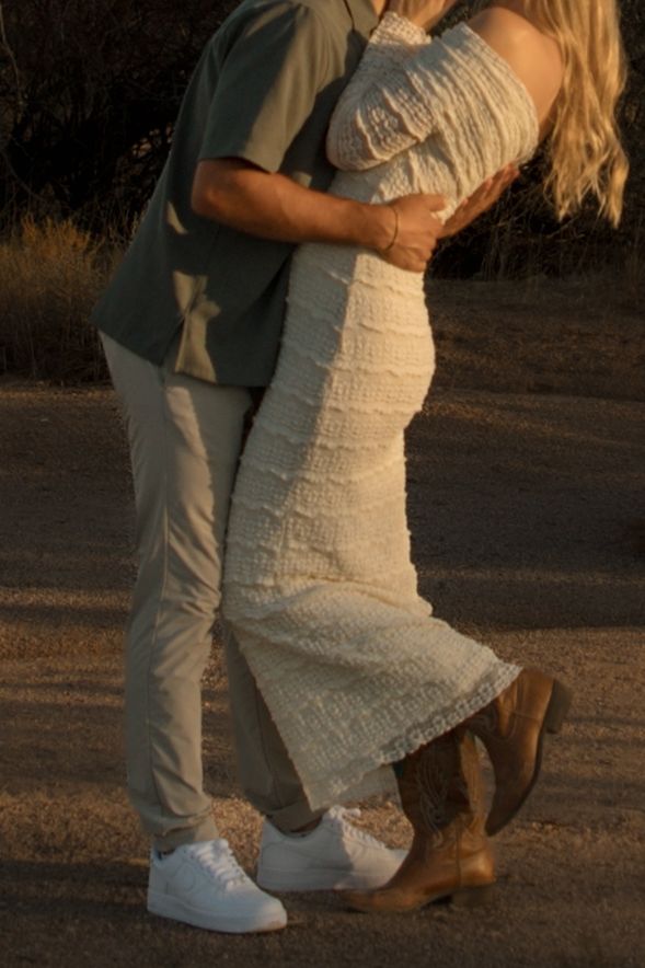 Couple embracing at golden hour, woman in white lace midi dress, man in sage green shirt and khaki pants outdoors.