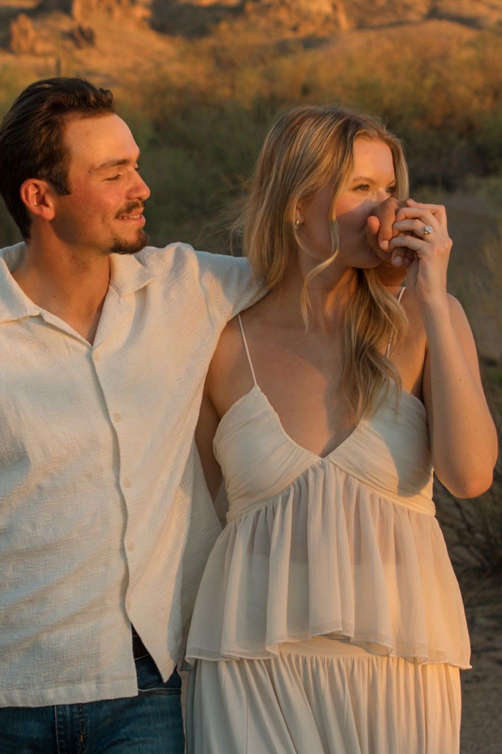 A couple in white attire share a romantic moment at golden hour in a desert landscape.