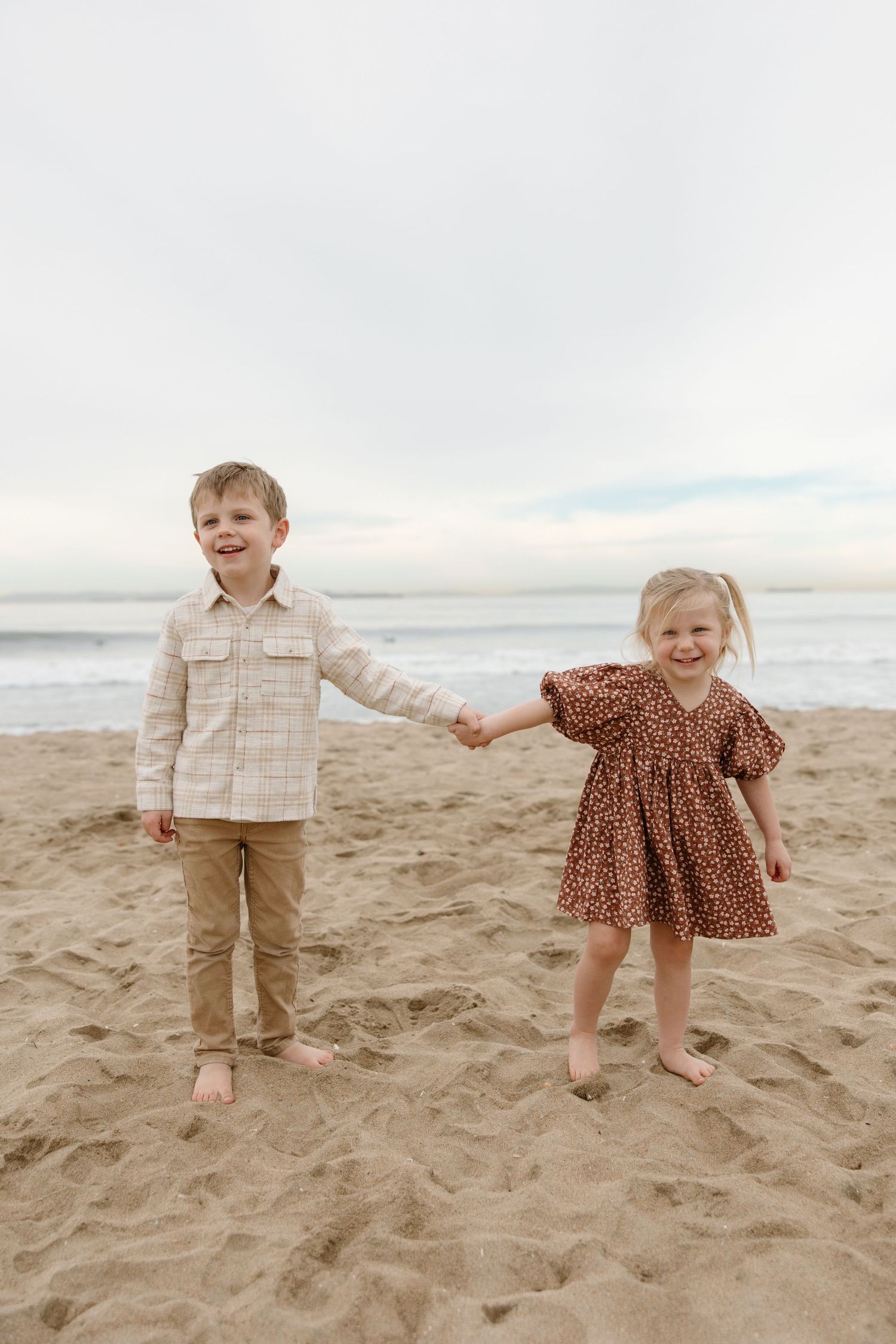Two young children holding hands and smiling on a sandy beach, boy in plaid shirt and girl in floral dress.