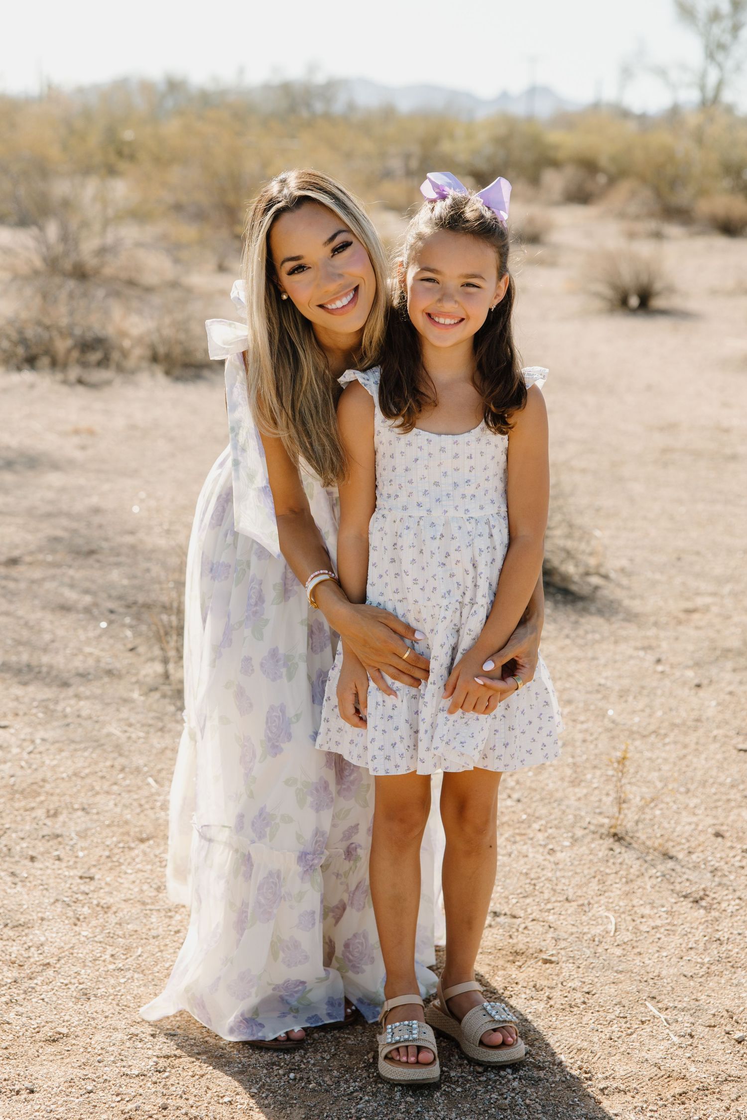 A smiling woman and young girl in matching floral dresses pose together in a desert landscape.
