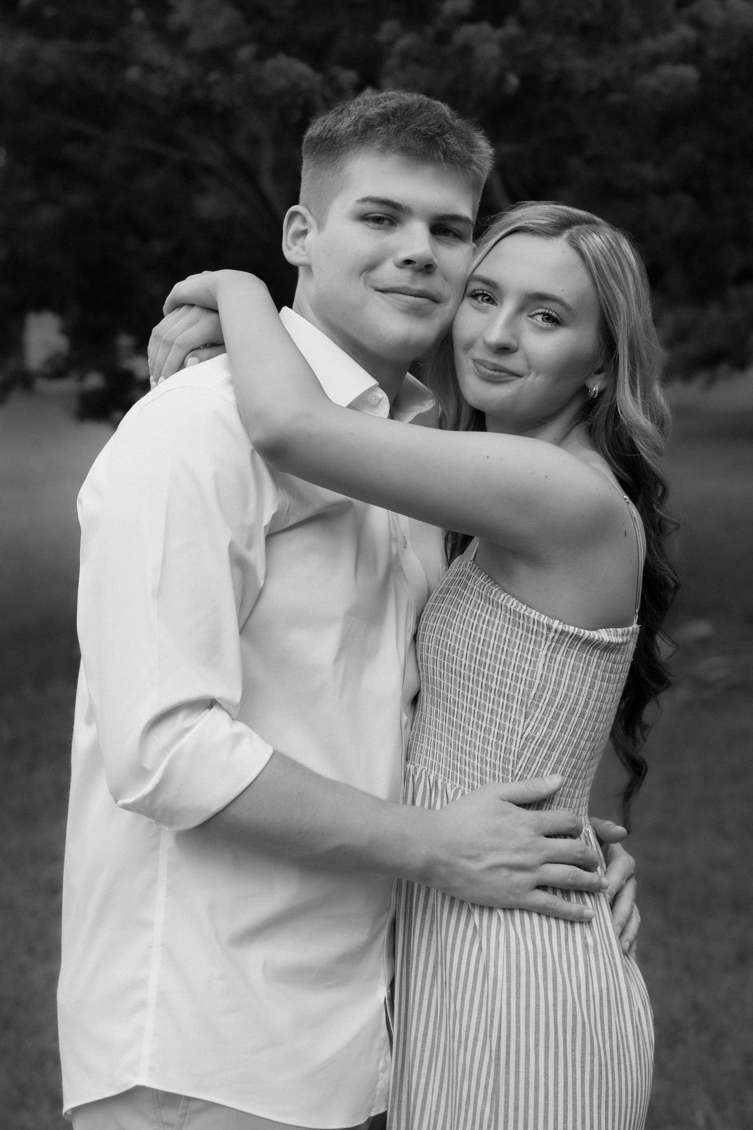 Black and white photo of a young couple embracing outdoors, smiling warmly in a park setting.