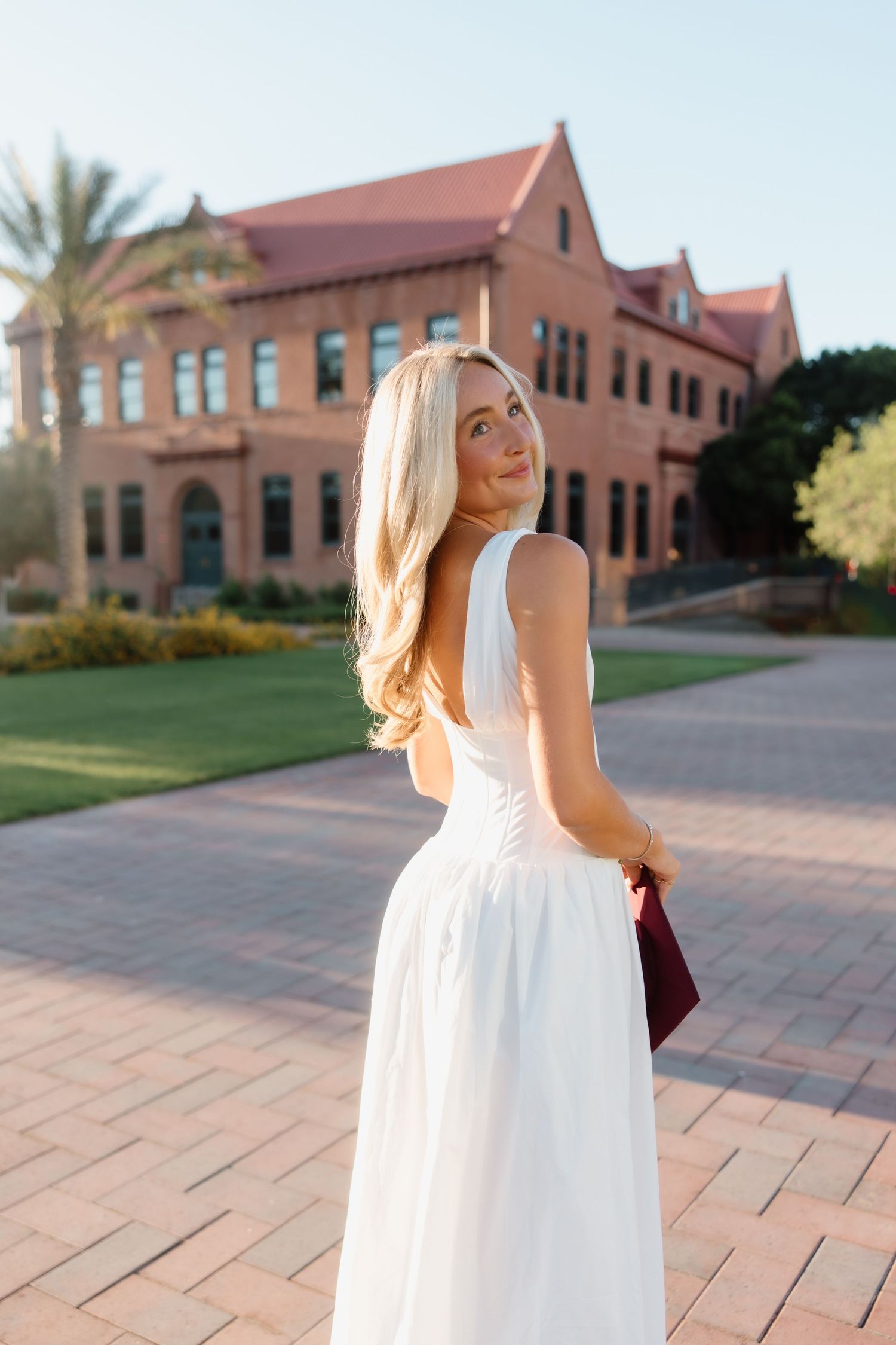 Blonde woman in white backless dress holding burgundy clutch on brick pathway outside red brick building.