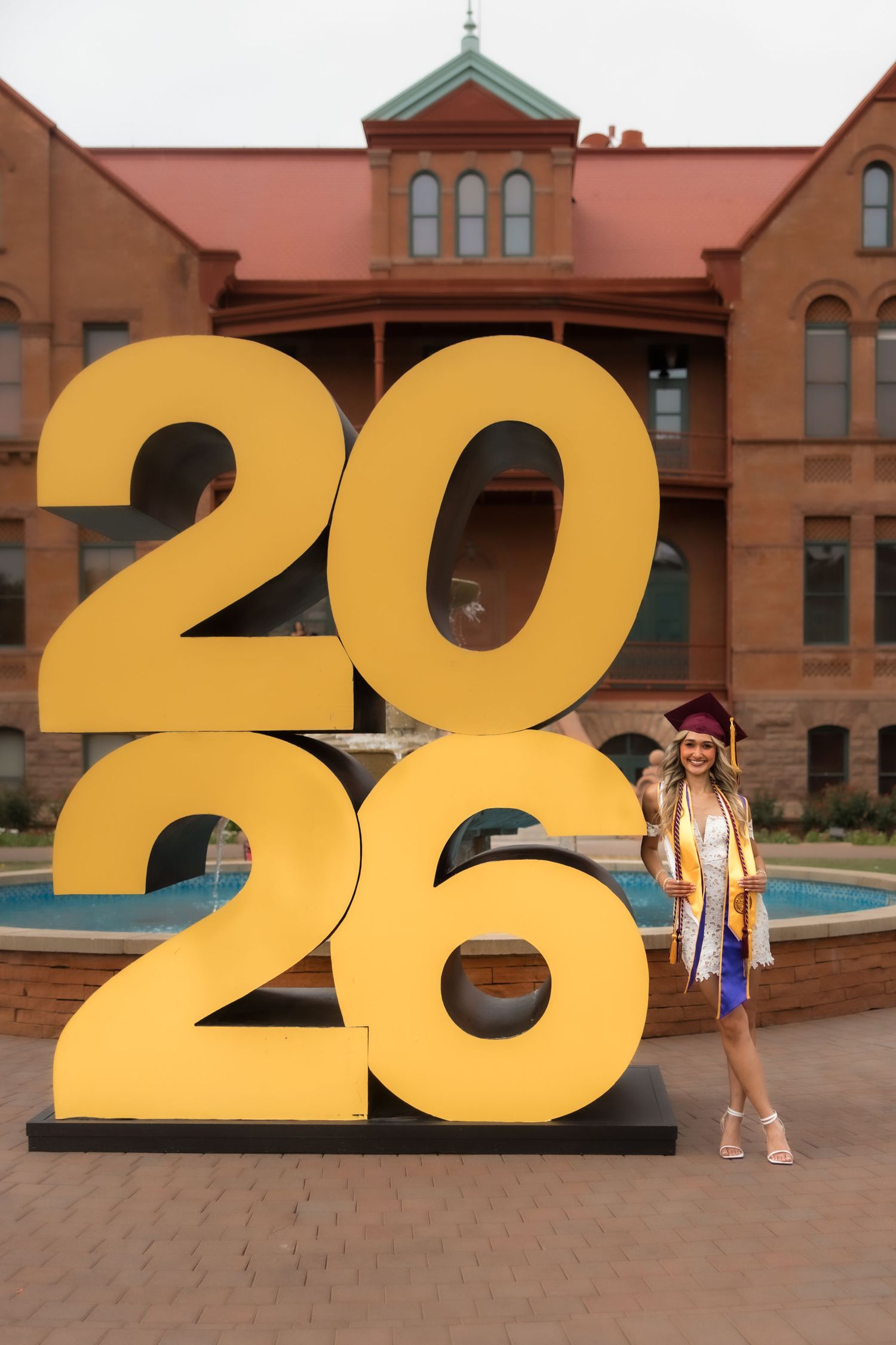 Graduate in cap and gown poses beside large gold 2026 sign on college campus with fountain and brick building behind.