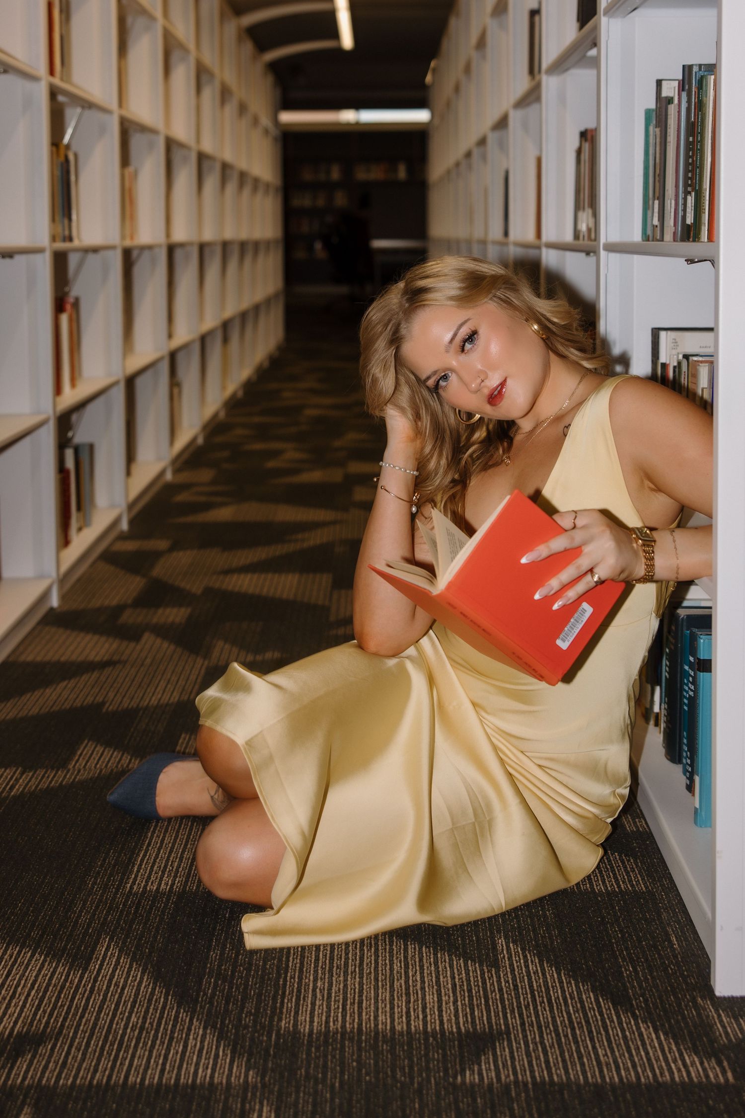 Woman in gold satin dress sitting on library floor reading a red book among white bookshelves.