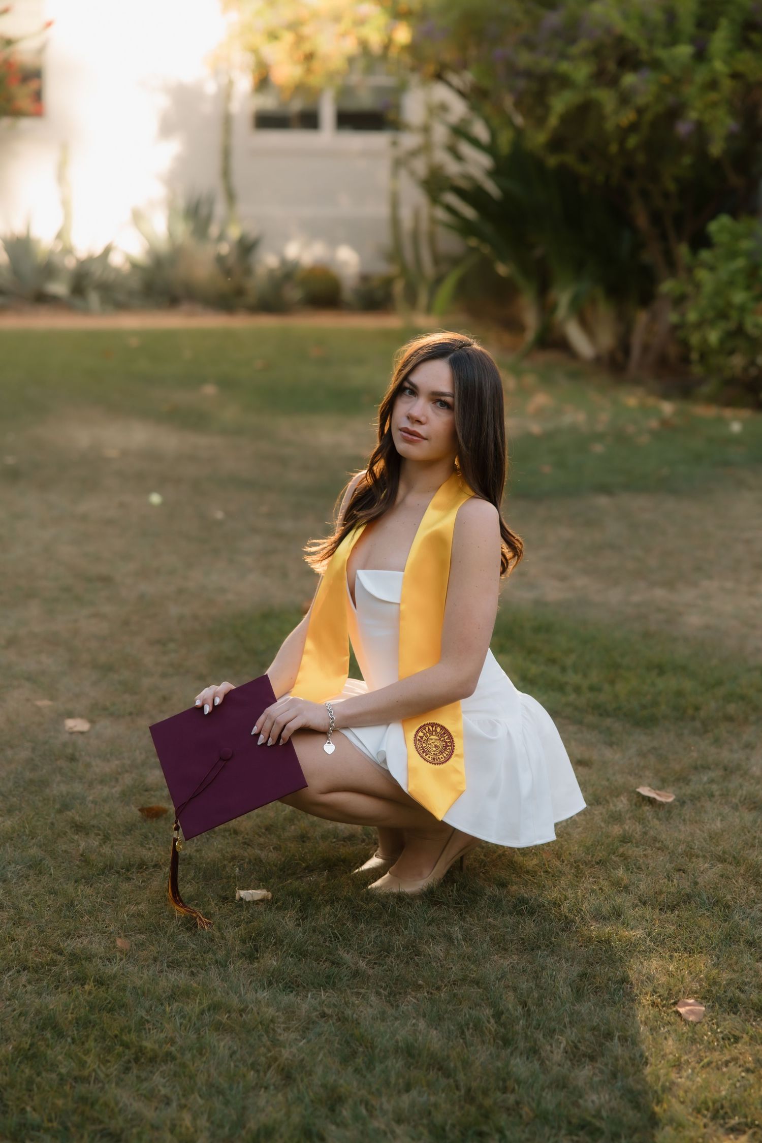 Young woman in white dress with gold graduation stole crouches on grass holding maroon cap.