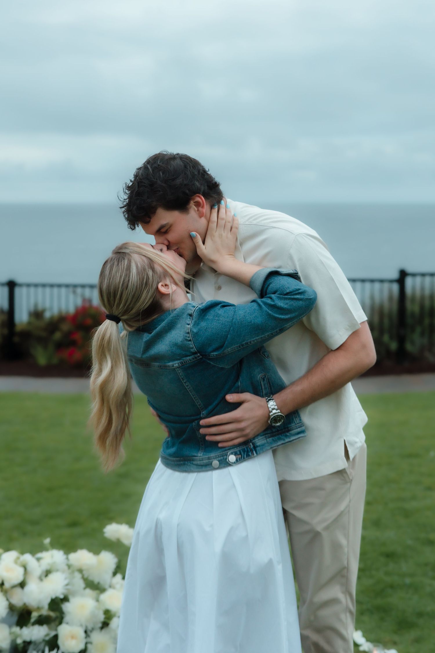 Couple sharing romantic kiss on garden lawn with ocean visible in the background.