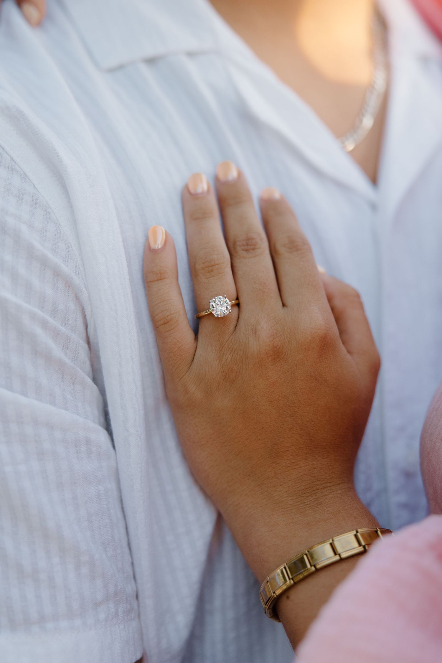 Close-up of a woman's hand displaying a diamond solitaire engagement ring and gold bracelet resting on a man's chest.