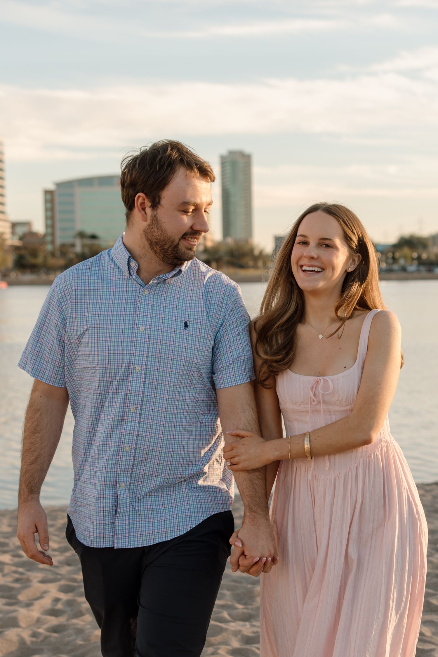 A couple walks hand-in-hand on a sandy beach at sunset, smiling, with a city skyline in the background.