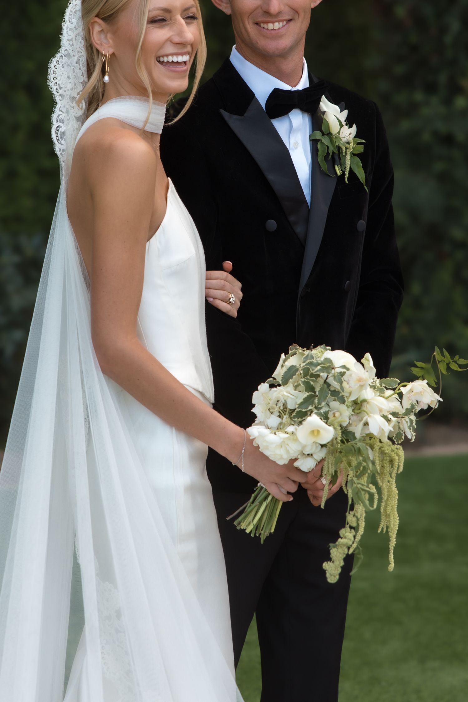 Laughing bride in white gown and veil holds white bouquet beside smiling groom in black tuxedo.