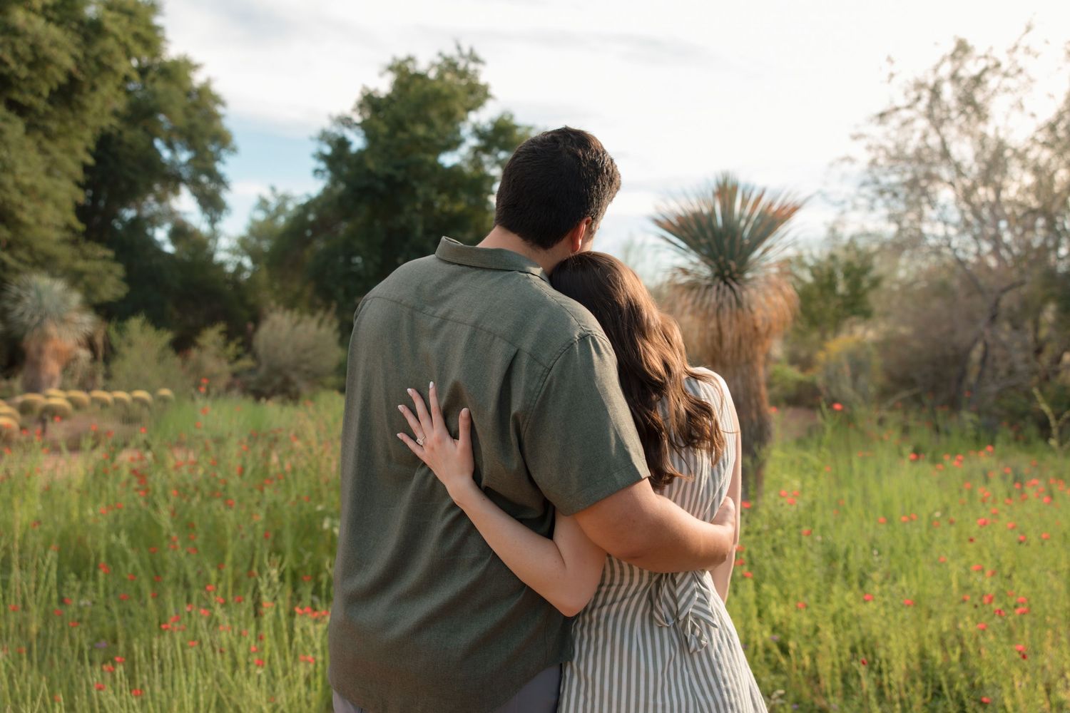 Couple walking arm in arm through a lush garden with wildflowers and green trees in warm sunlight.
