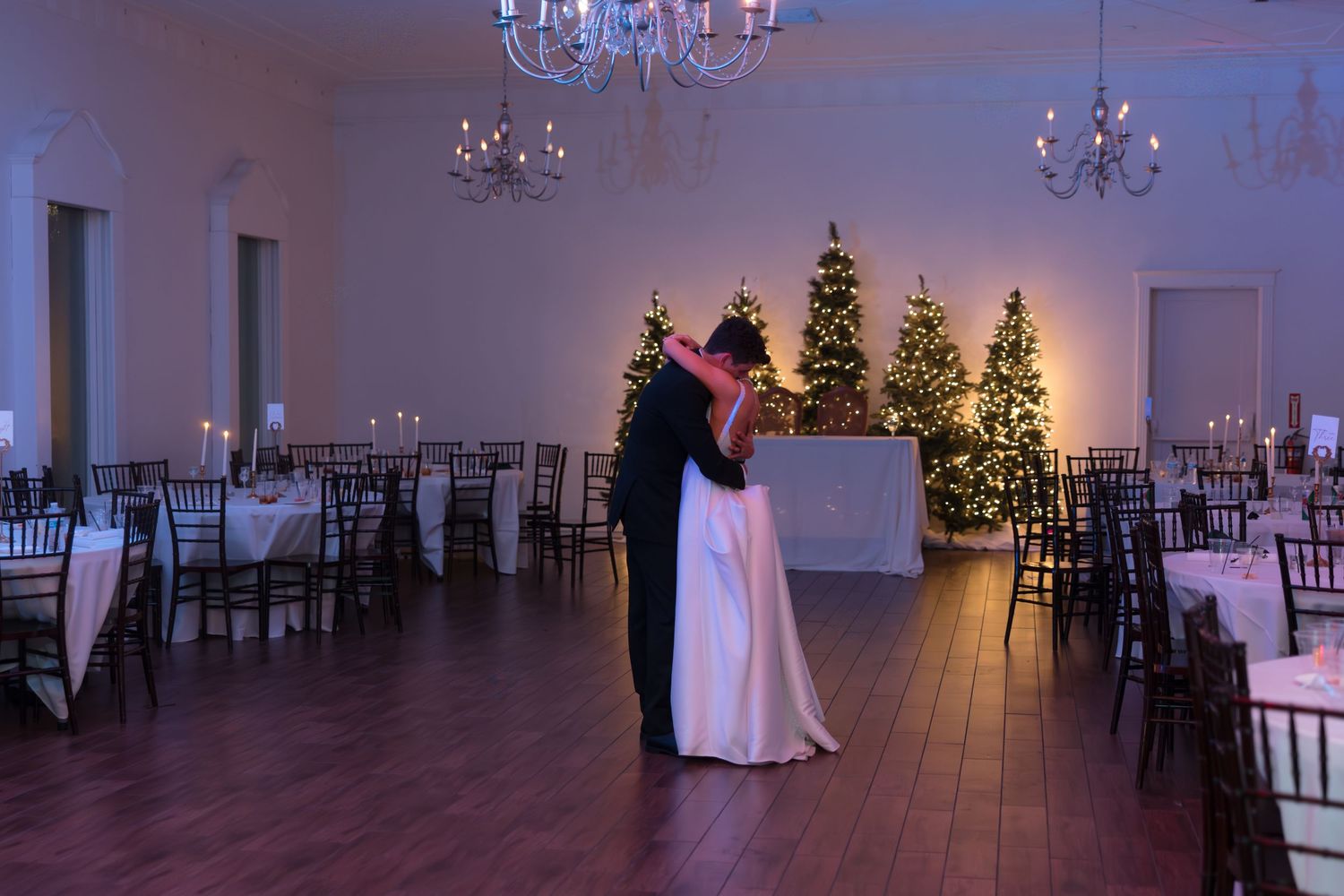 Bride and groom share romantic first dance in elegant ballroom adorned with glowing Christmas trees and chandeliers.