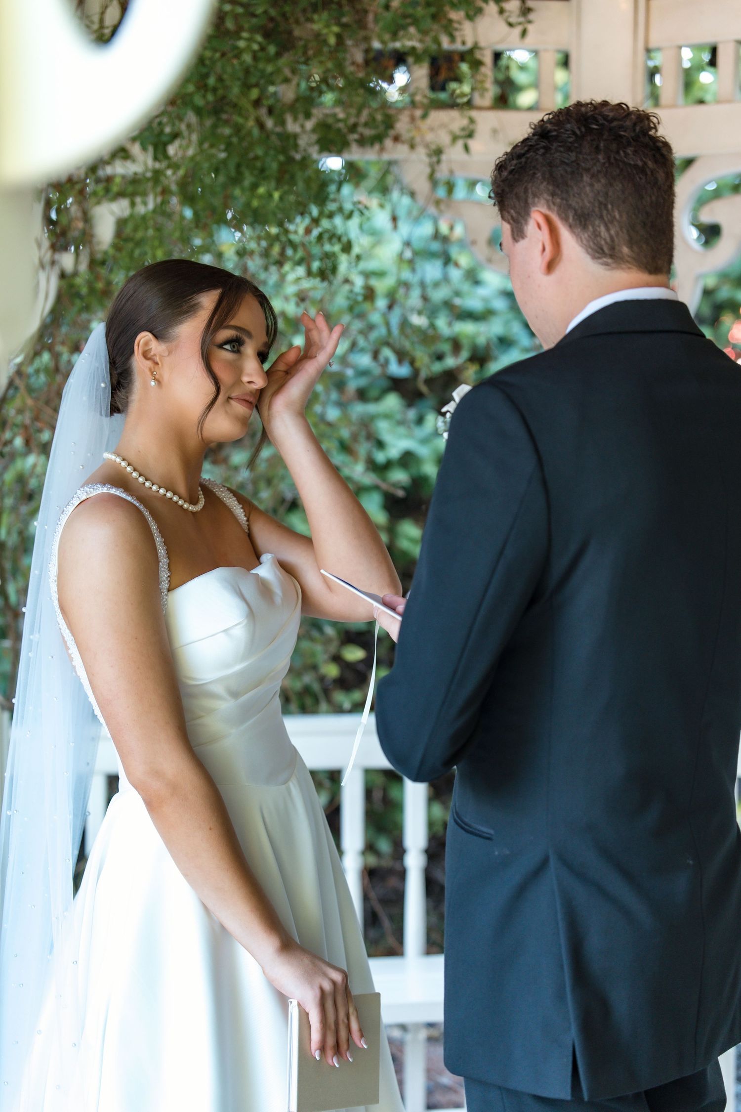 Emotional bride wiping tears during outdoor wedding ceremony while groom reads vows at garden altar.