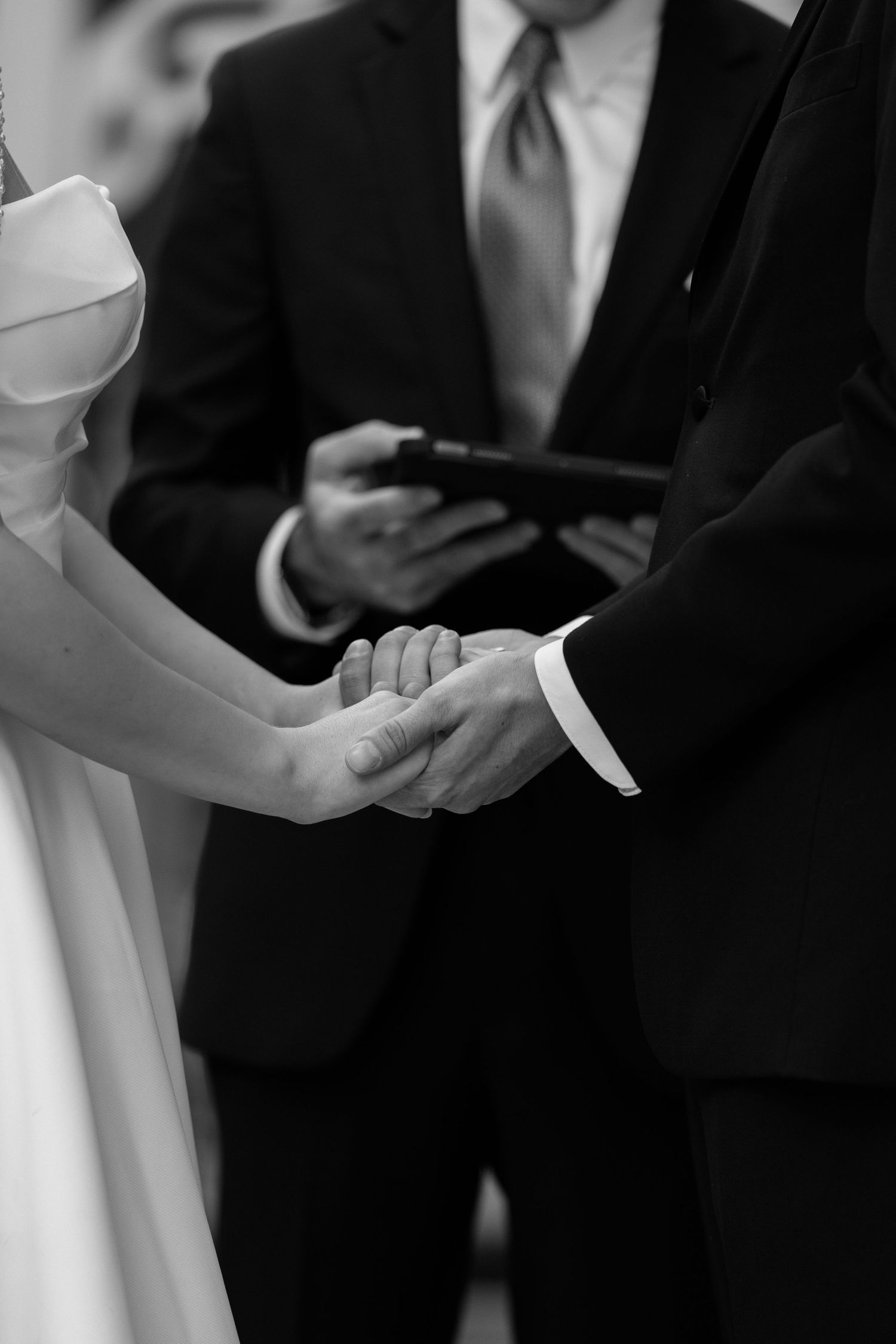Black and white photo of bride and groom holding hands during wedding ring exchange ceremony.