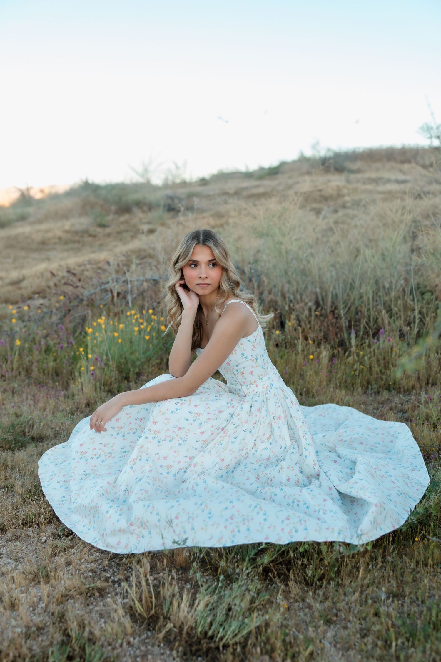 Young woman in flowing white floral dress seated in grassy hillside field for elegant senior portraits.