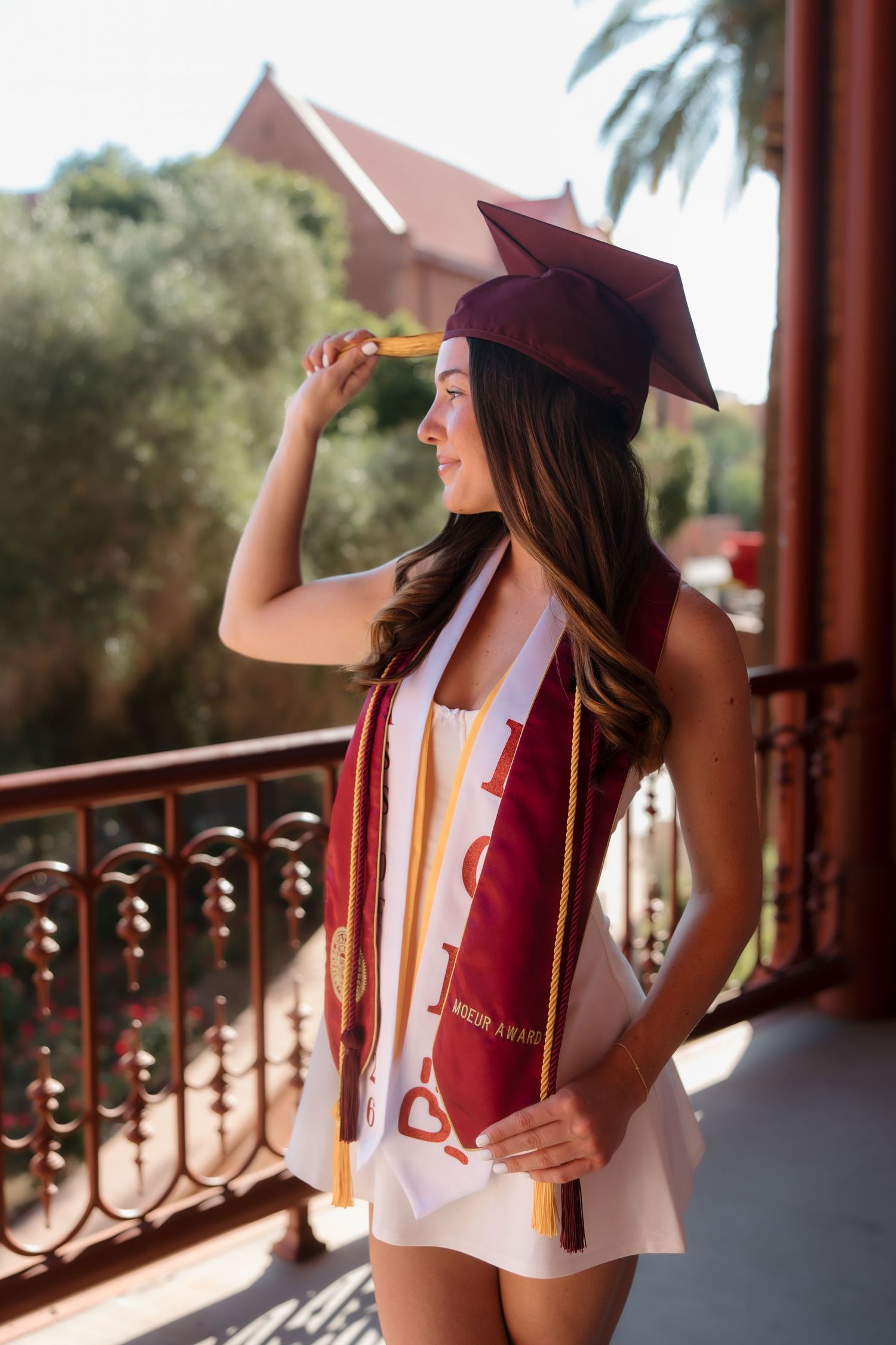 Female graduate in maroon cap and white dress with honor stoles posing on sunlit campus balcony.