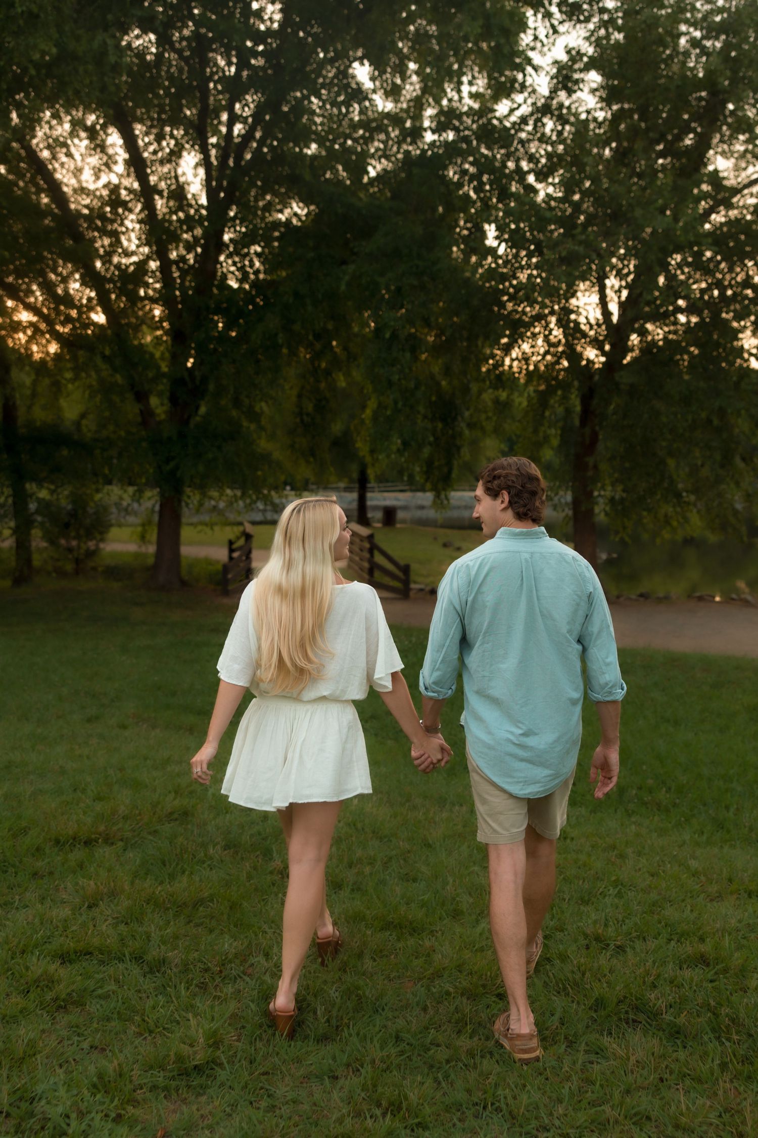Couple walking hand in hand away from camera through a lush green park at golden hour sunset.