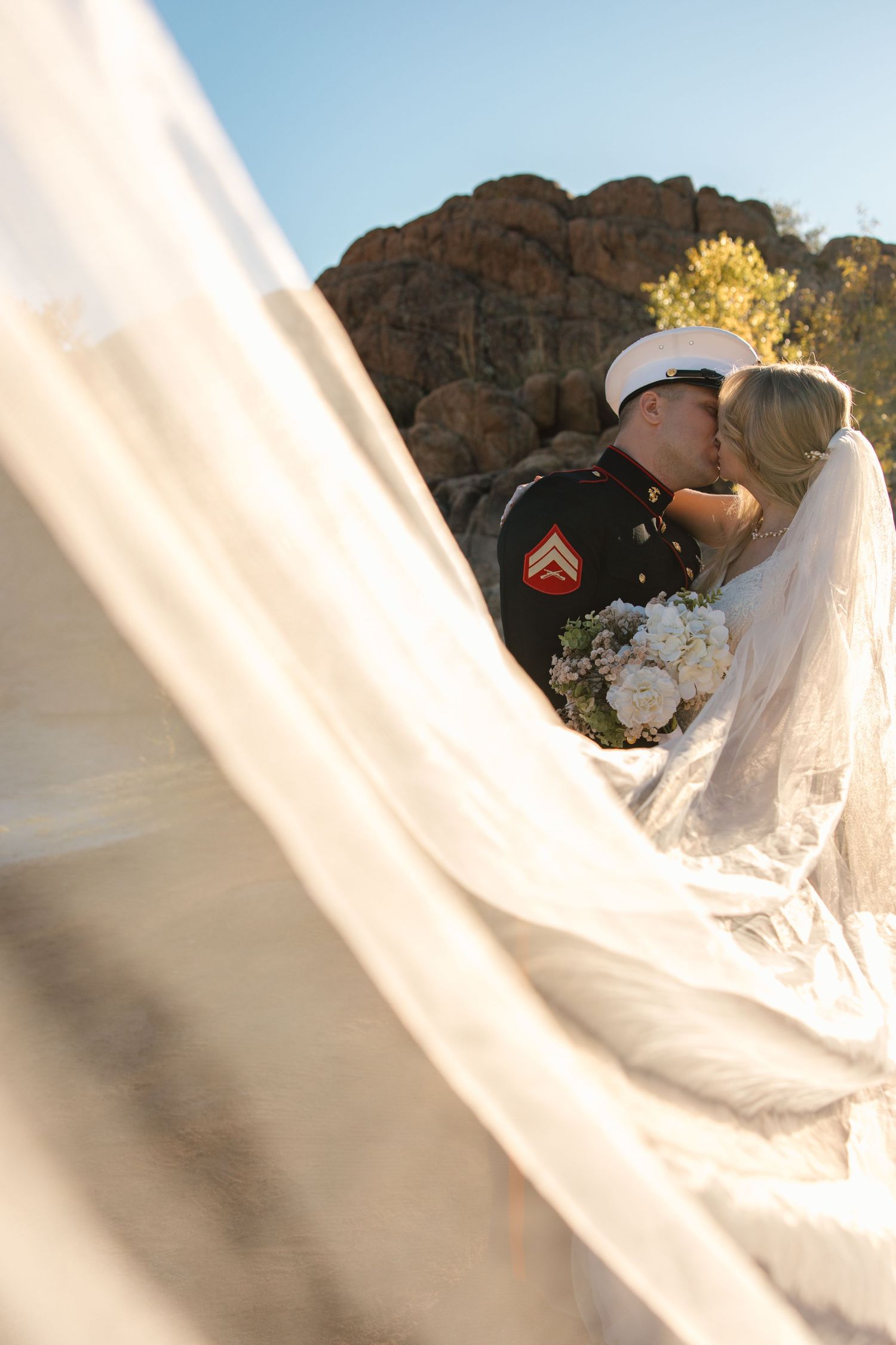 Marine groom kisses bride framed by flowing veil with rocky landscape in golden sunlight background.