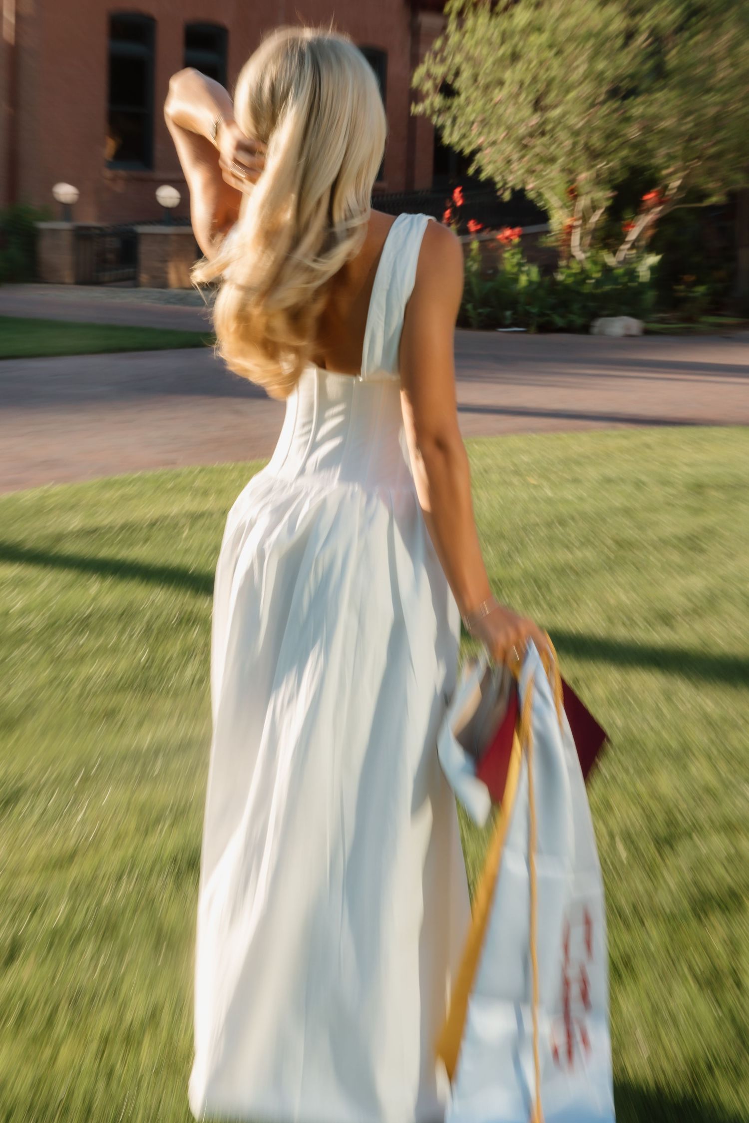Blonde woman in elegant white backless gown walks across green campus lawn holding graduation cap and stole.