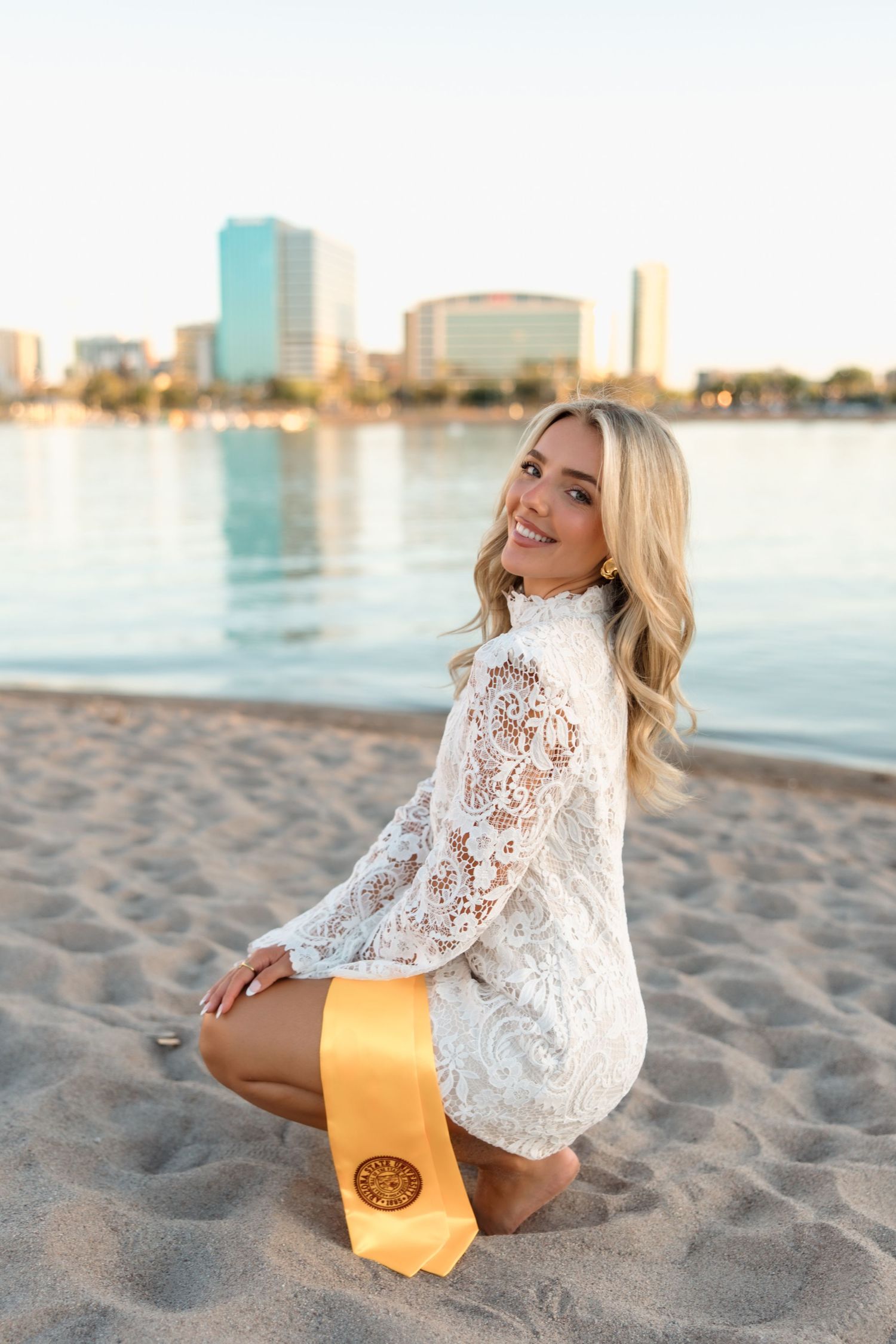 Smiling blonde graduate in white lace dress sits on beach with gold stole and city skyline.