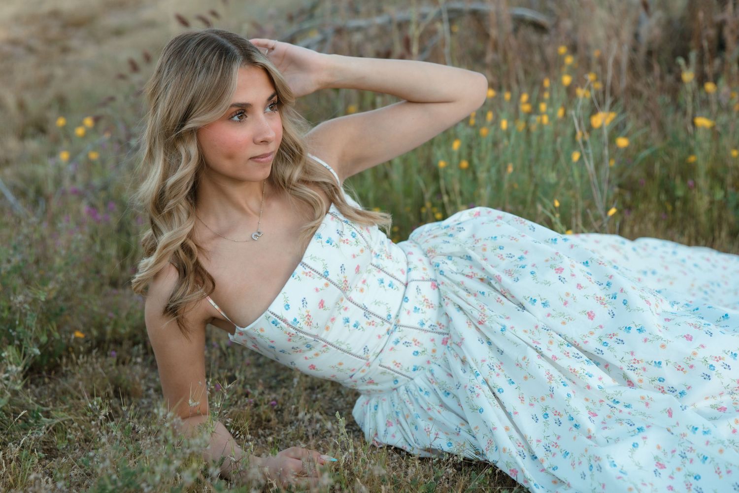 Young woman in floral sundress reclining in a wildflower meadow, posing gracefully with hand in blonde hair.