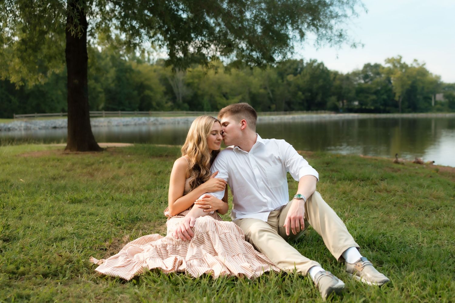 Couple sitting on grass by a lake, sharing a romantic moment under a tree in a lush park setting.