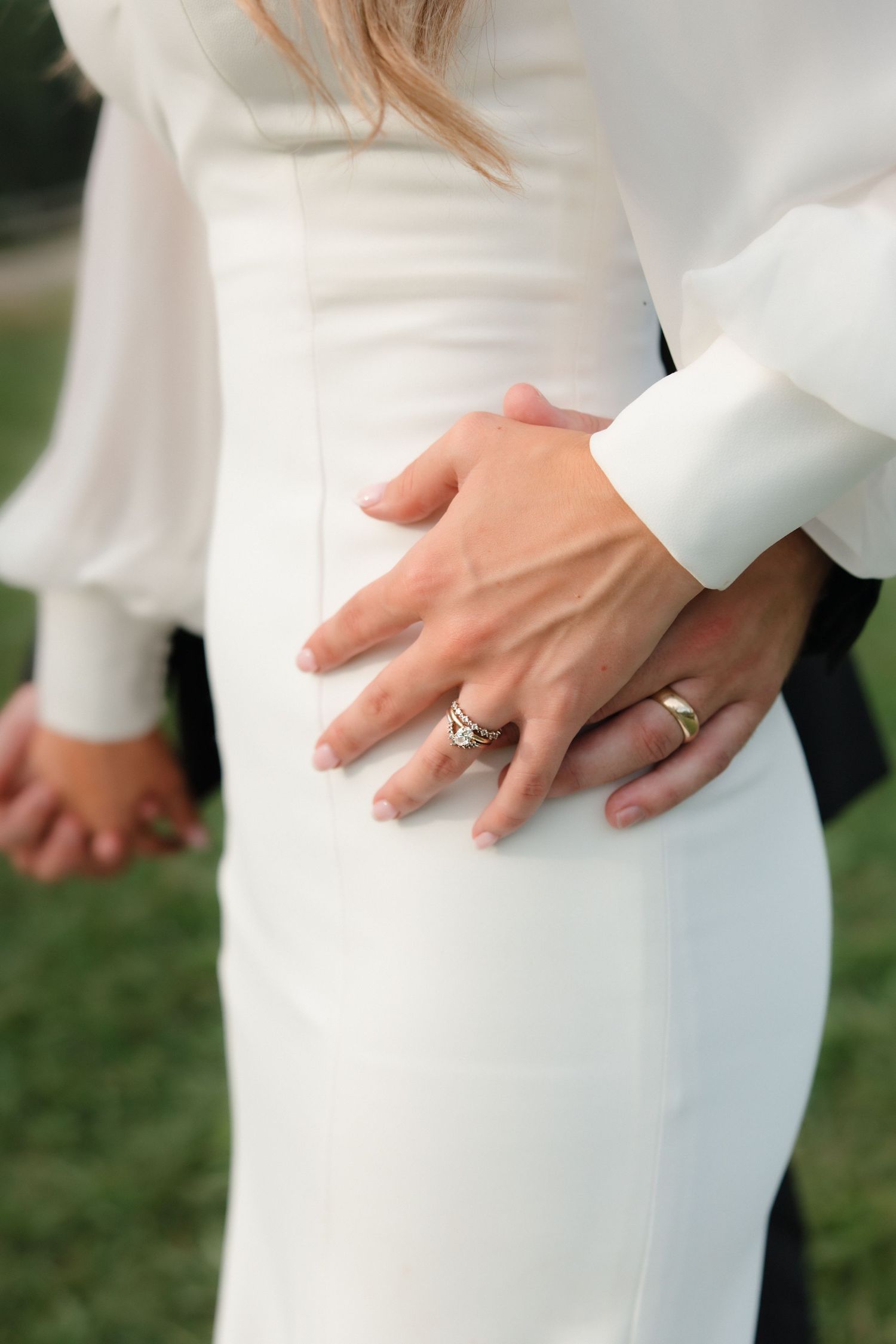 Close-up of bride and groom hands showing wedding and engagement rings on white bridal gown.