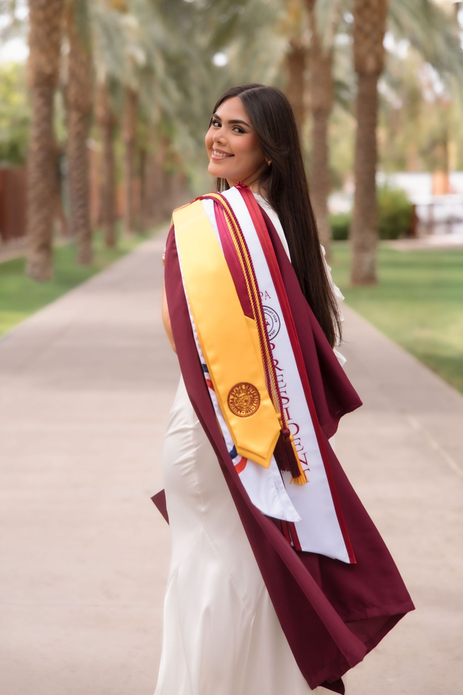Smiling graduate in maroon cap and gown with gold sash poses outdoors among palm trees on graduation day.