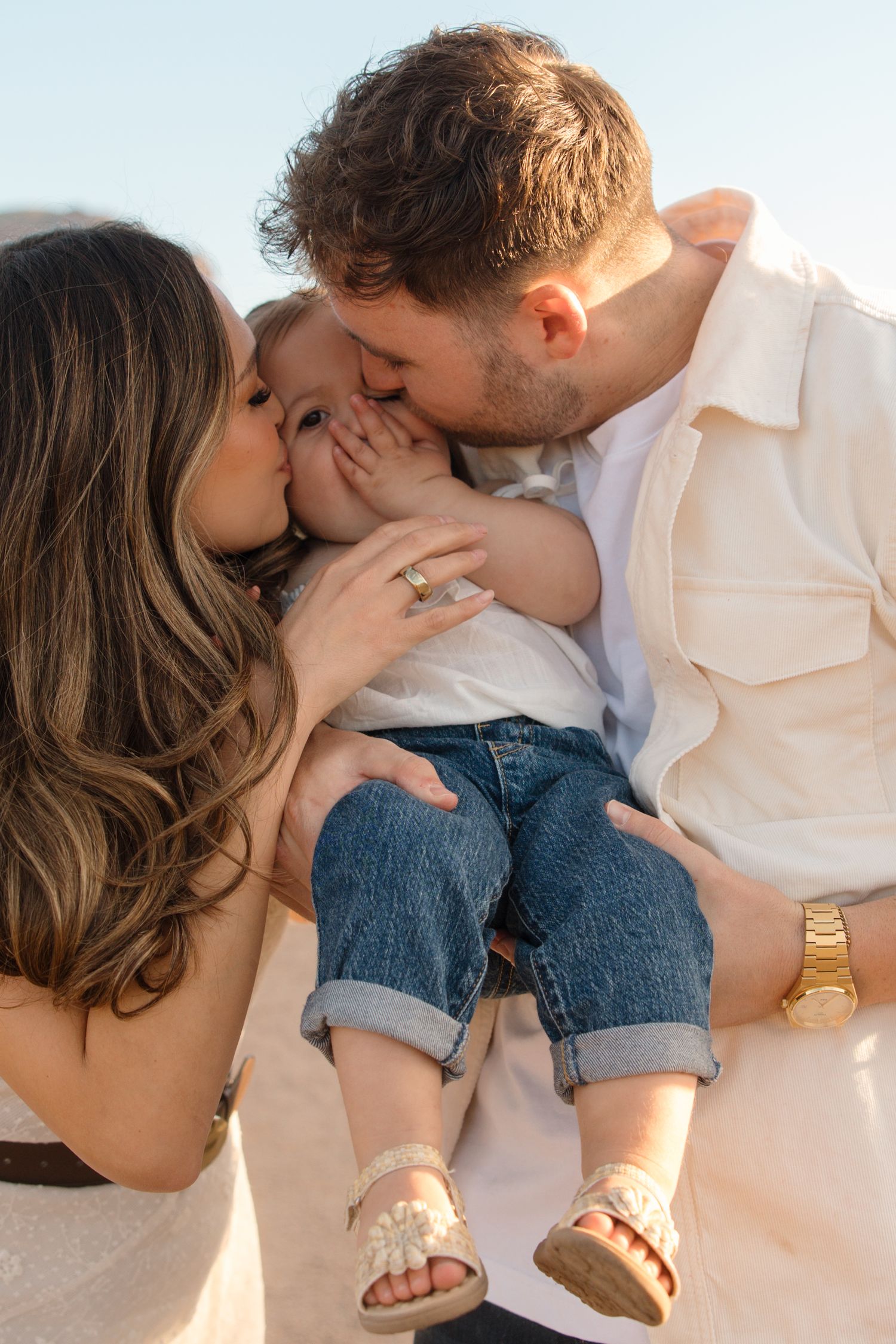 Loving parents kissing their toddler daughter simultaneously in a sunny outdoor setting.