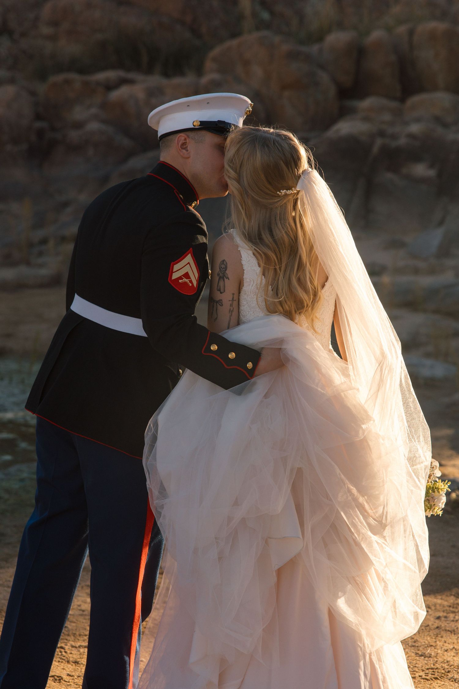 Marine groom in dress uniform shares first kiss with bride in flowing white gown at desert wedding.