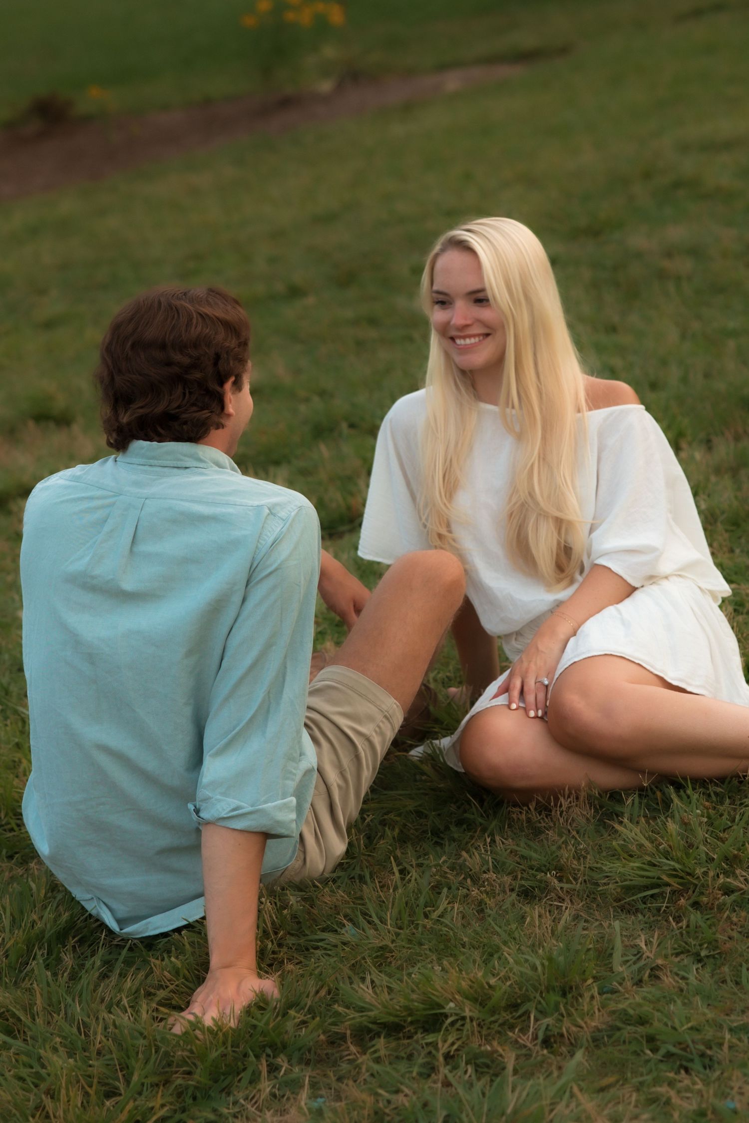 Smiling blonde woman in white dress sitting on grass chatting with her partner outdoors.