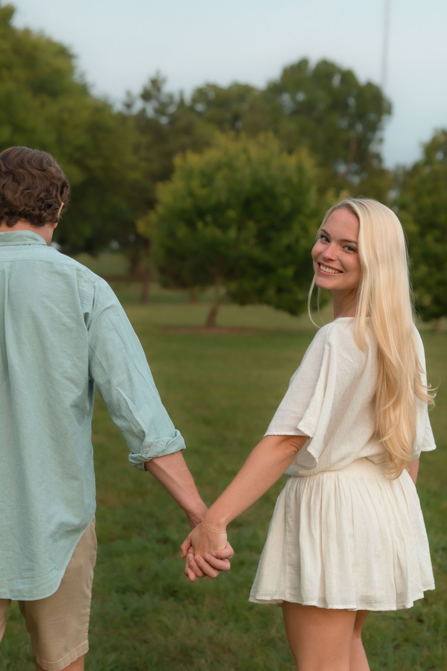 Smiling blonde woman glances back while holding hands with her partner in a park.