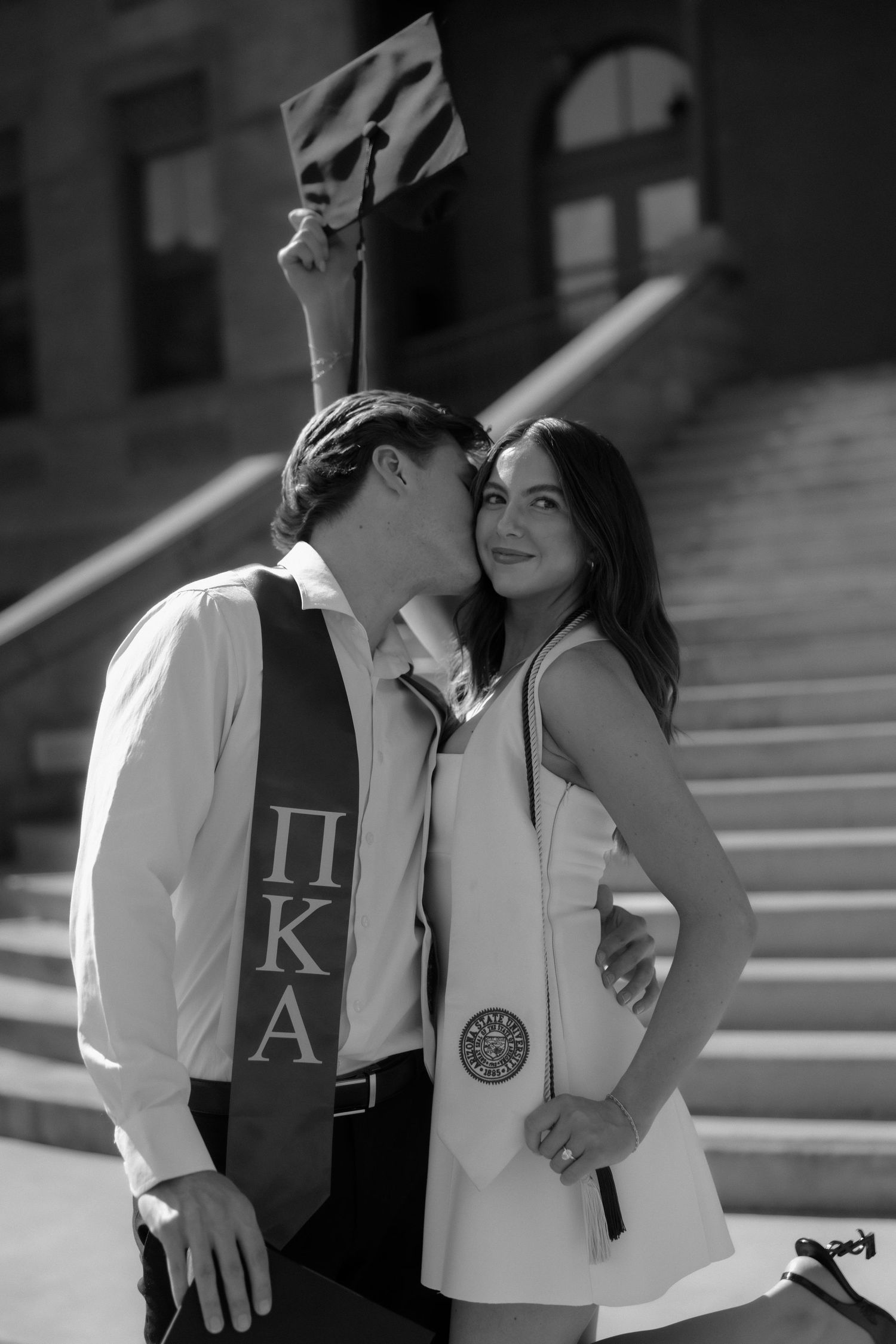 Black and white photo of graduate in Pi Kappa Alpha stole kissing woman holding maroon cap on steps.