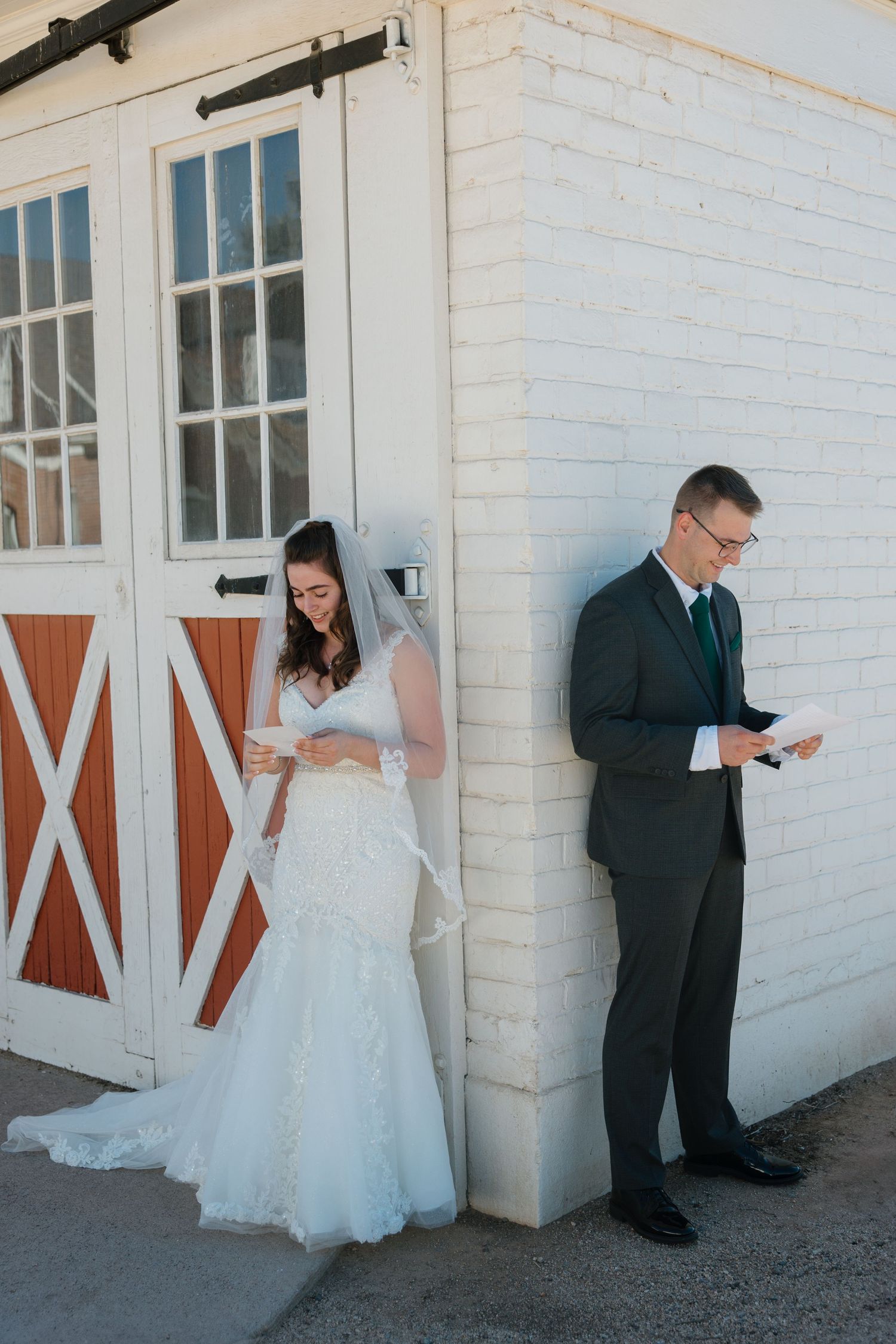 Bride and groom stand back-to-back reading heartfelt letters before their wedding ceremony outside a white barn.