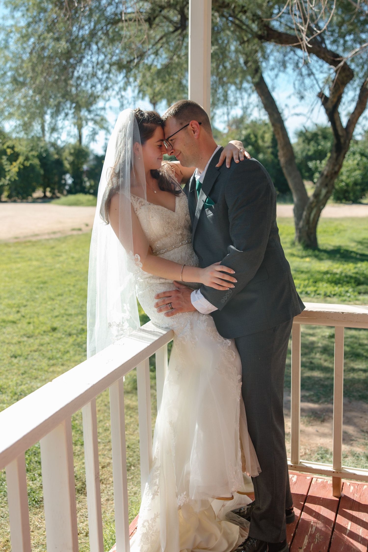 Bride and groom share a romantic moment on a white porch railing surrounded by lush green trees.