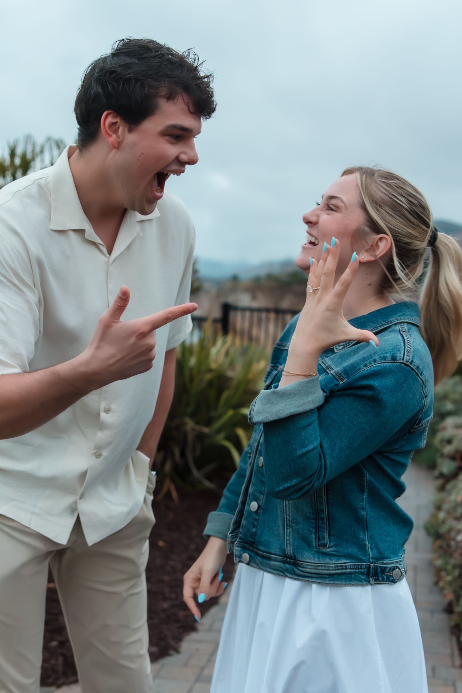 Excited young couple laughing outdoors, man in white shirt pointing, woman in denim jacket touching her face.