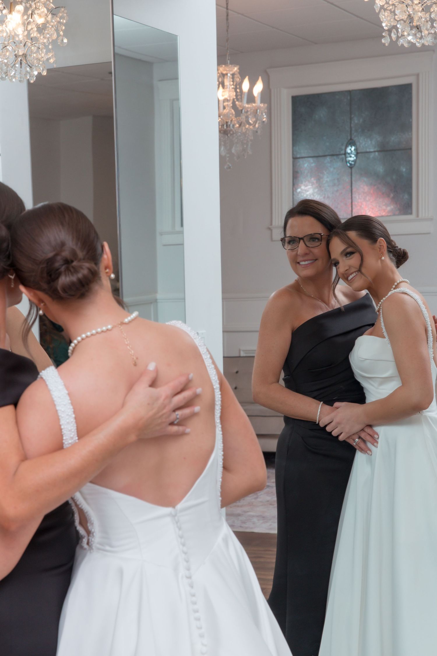 Bride in backless wedding gown embracing woman in black dress while bridesmaids look on in mirror.
