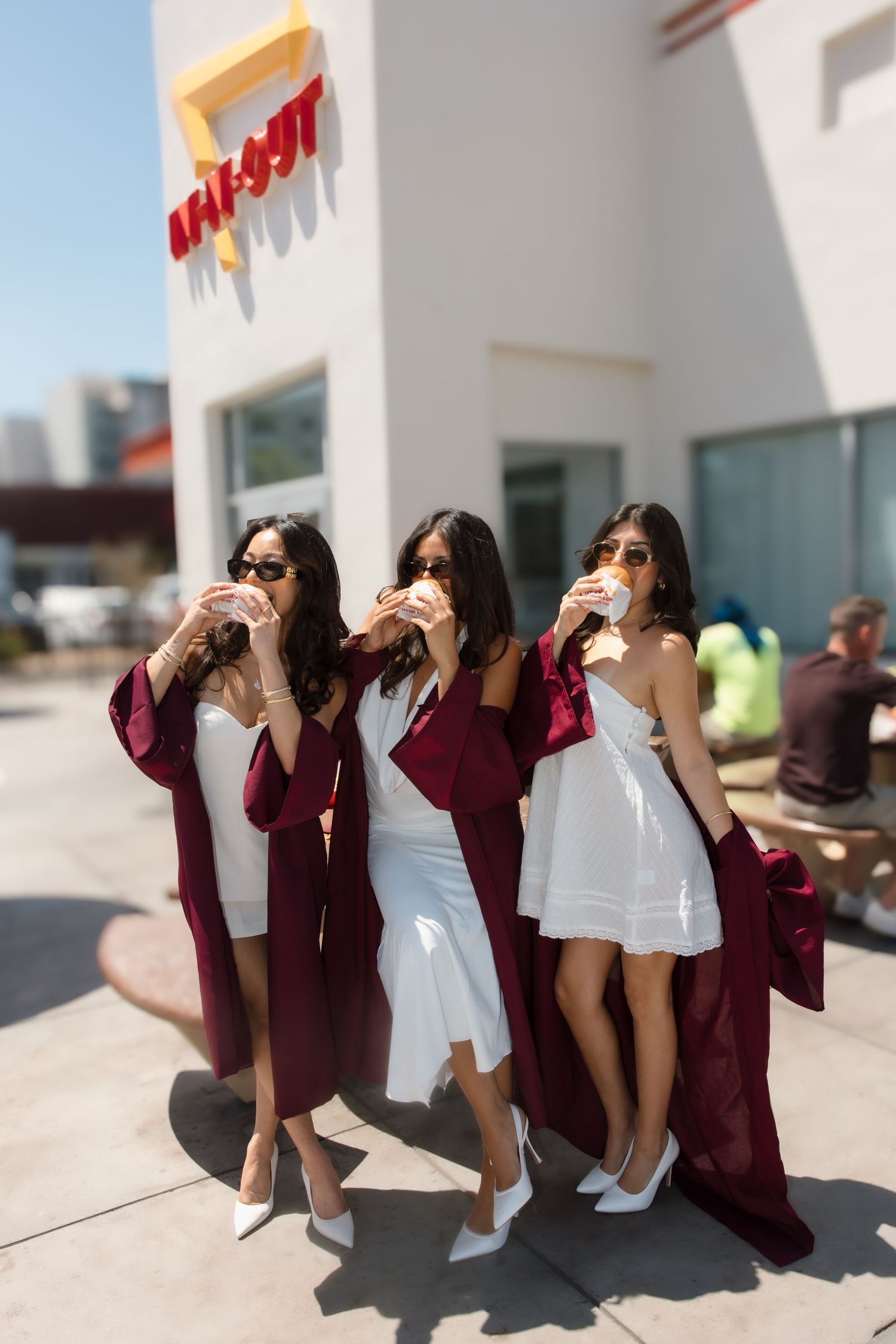 Three female graduates in burgundy robes and sunglasses eat burgers outside In-N-Out Burger.