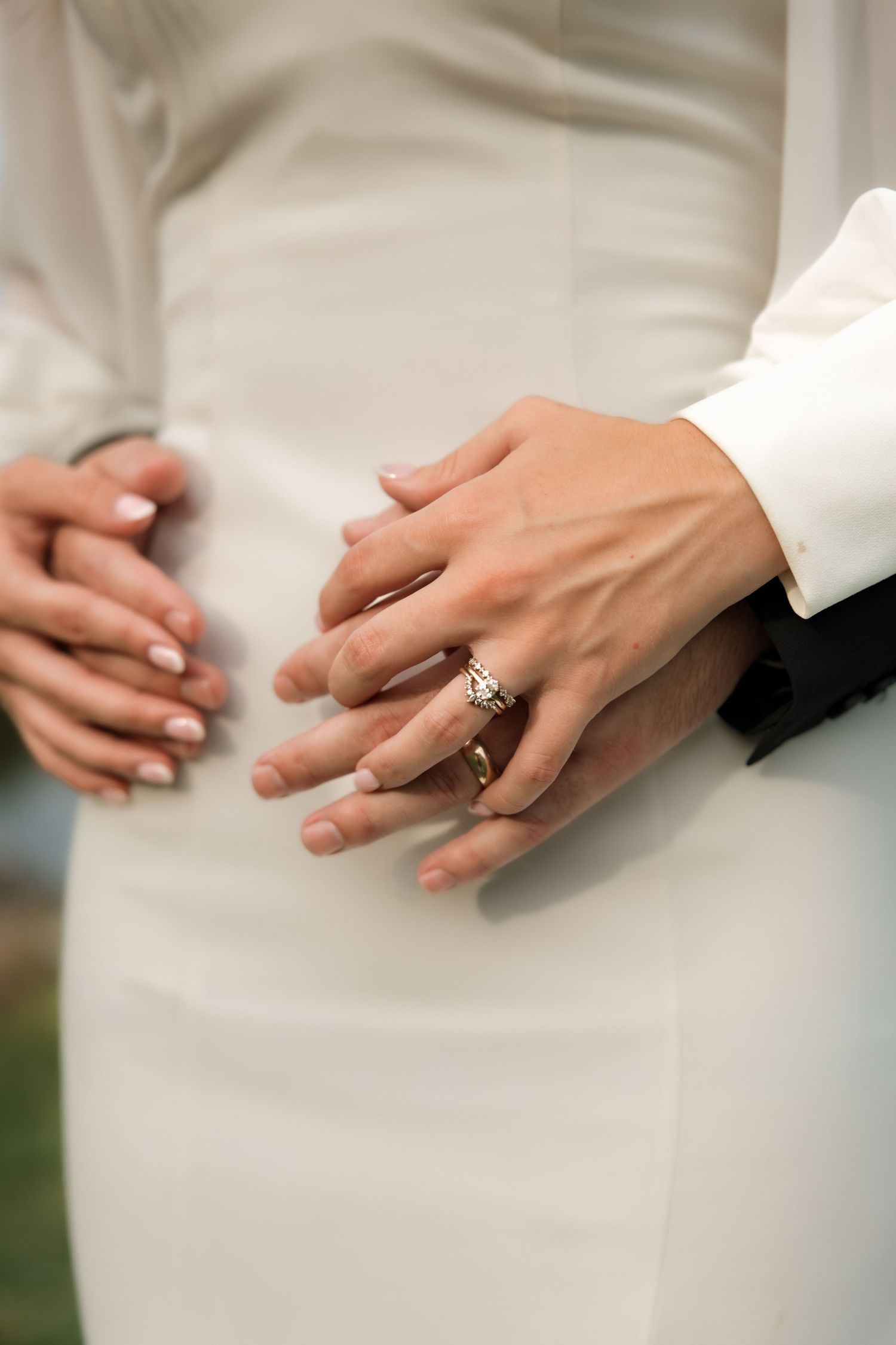 Close-up of newlyweds hands with wedding rings, bride in white dress, groom in black suit.