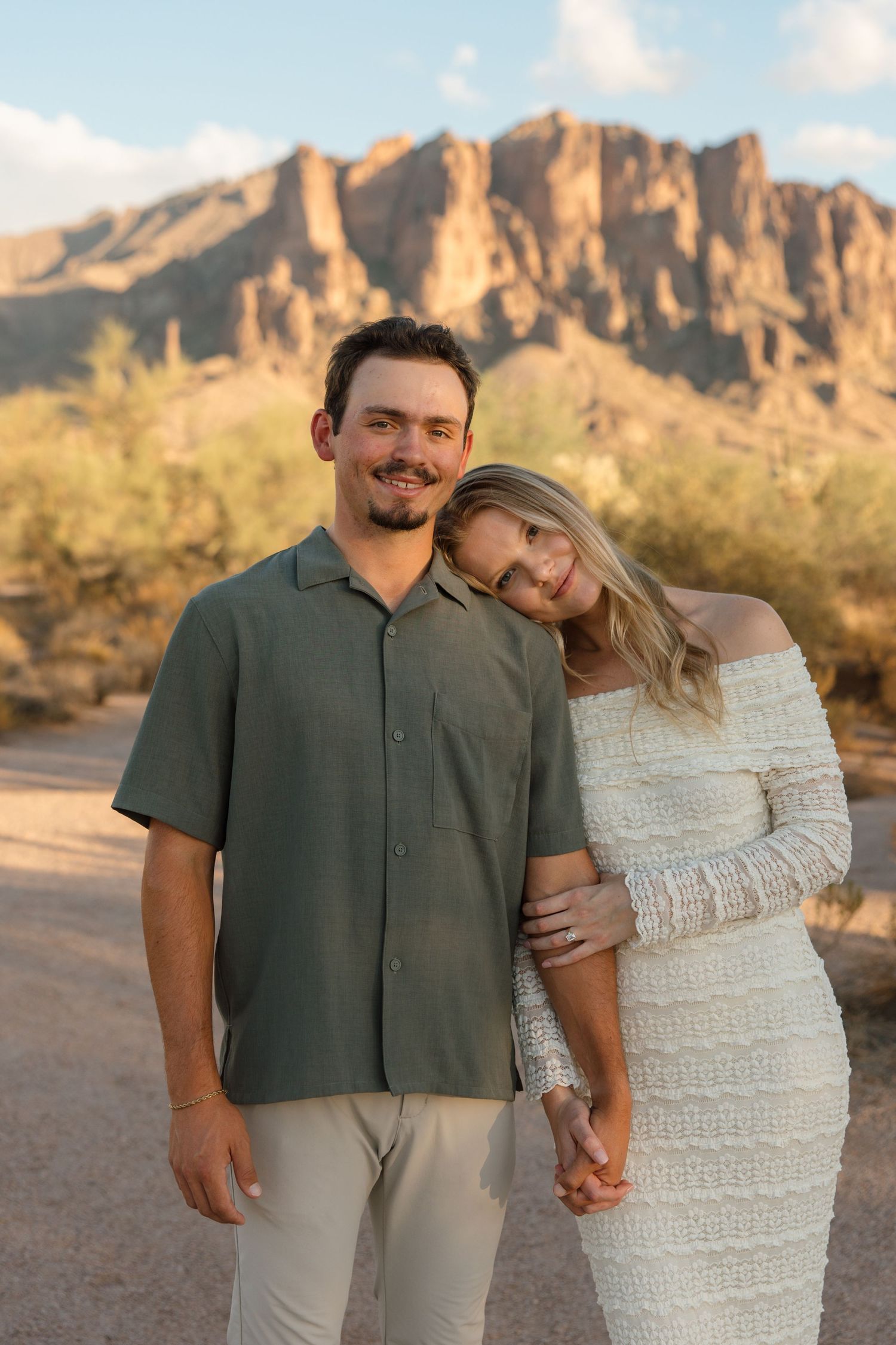 Couple posing in Arizona desert with Superstition Mountains backdrop, woman in white dress leaning on man in green shirt.