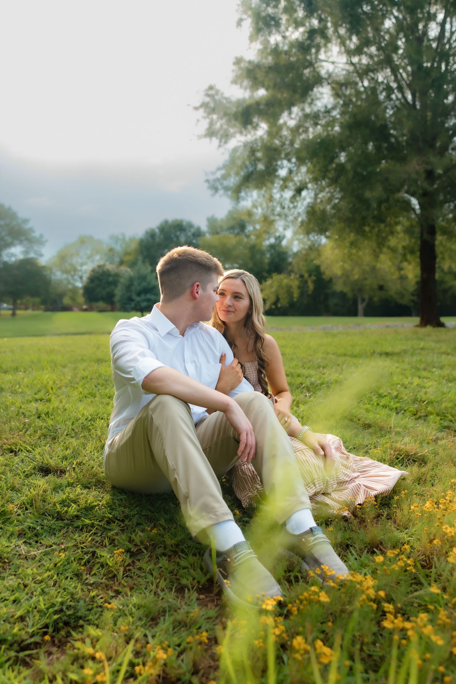 A couple sits together on green grass in a park, the woman in a flowy dress gazing at her partner on a sunny day.