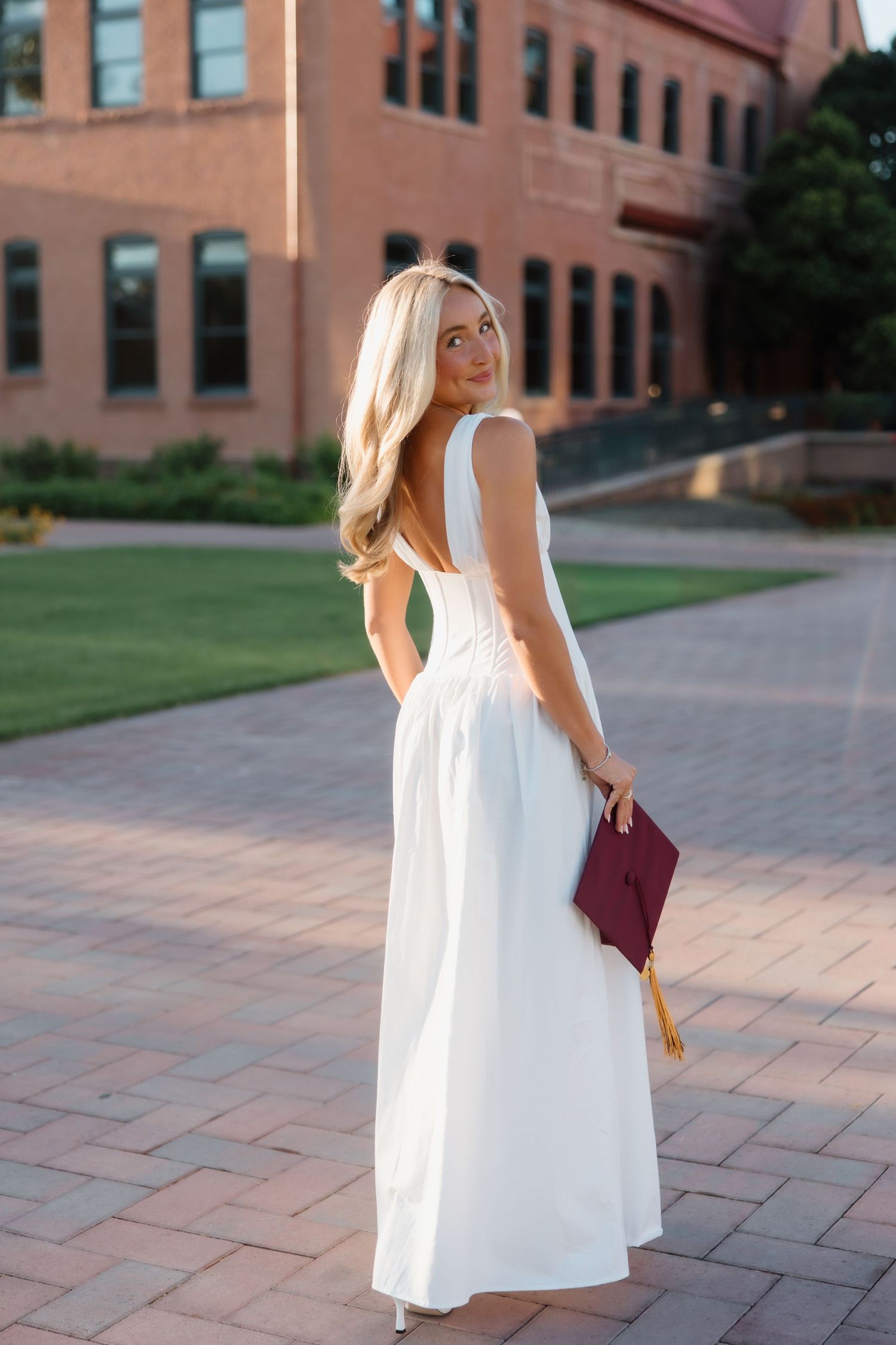 Blonde woman in elegant white backless dress holding maroon graduation cap on campus.