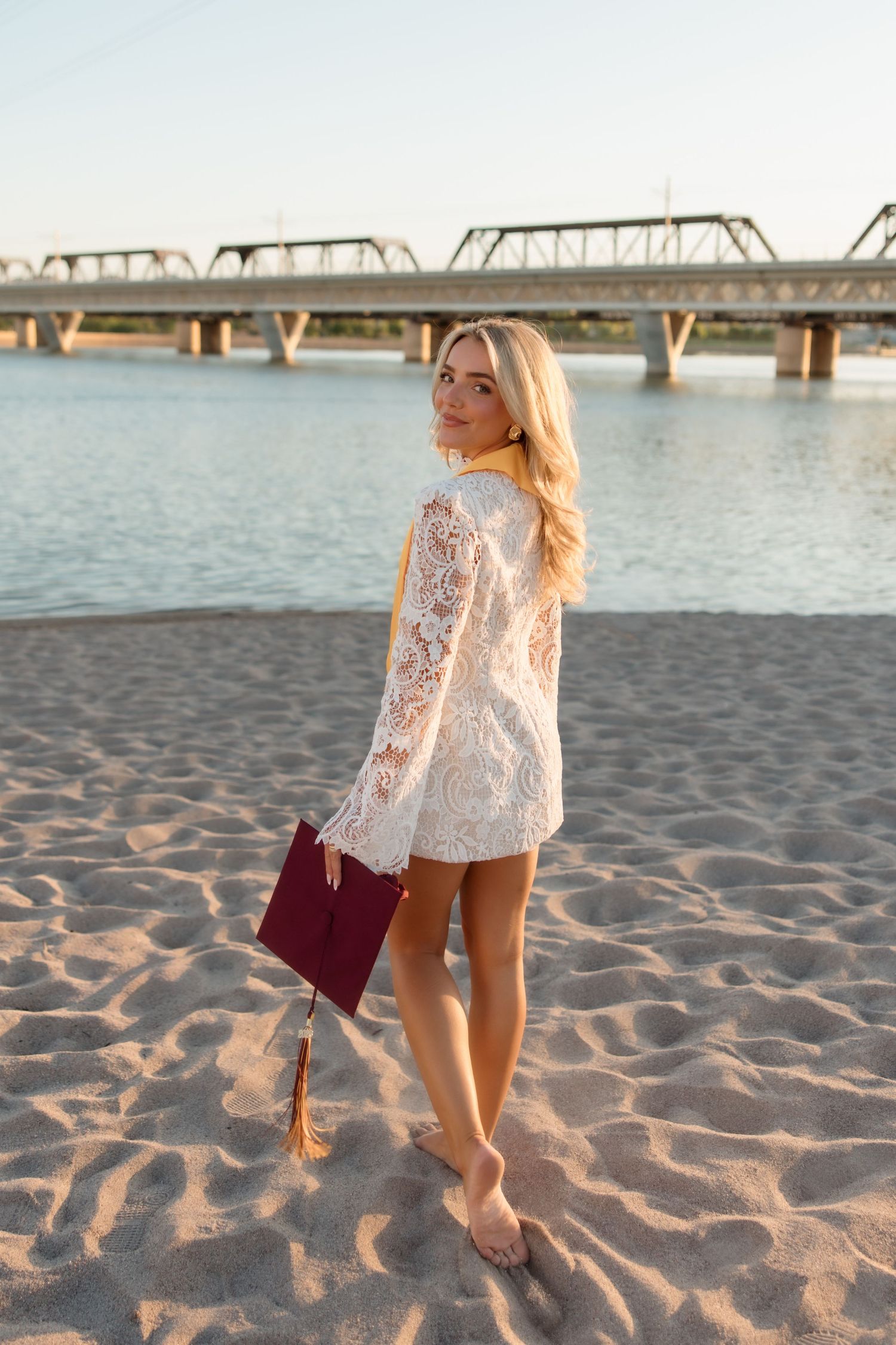 Blonde woman in white lace dress holding burgundy clutch on sandy beach with bridge and river backdrop.