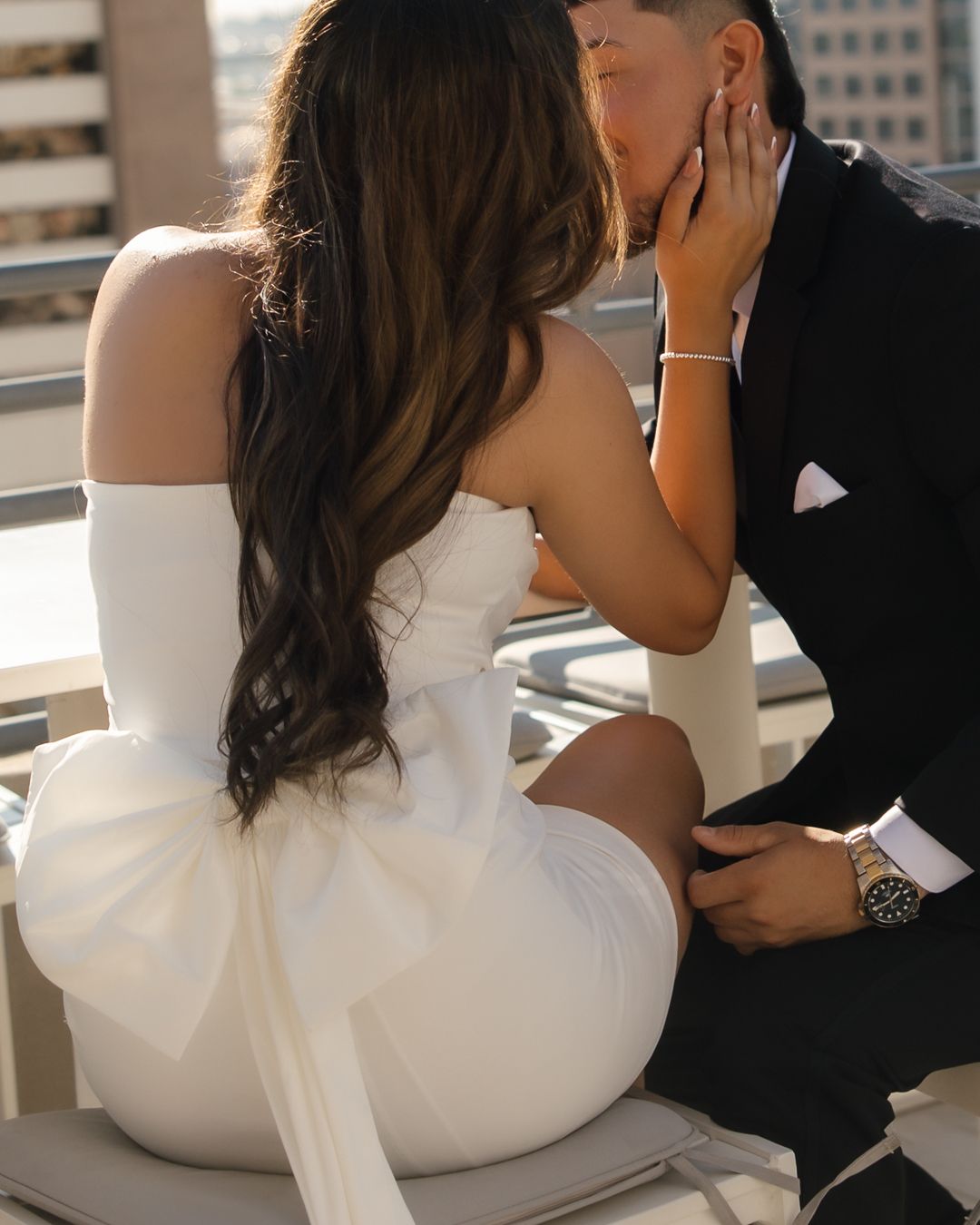Couple sharing a romantic kiss, bride in white off-shoulder bow dress and groom in black suit on rooftop.